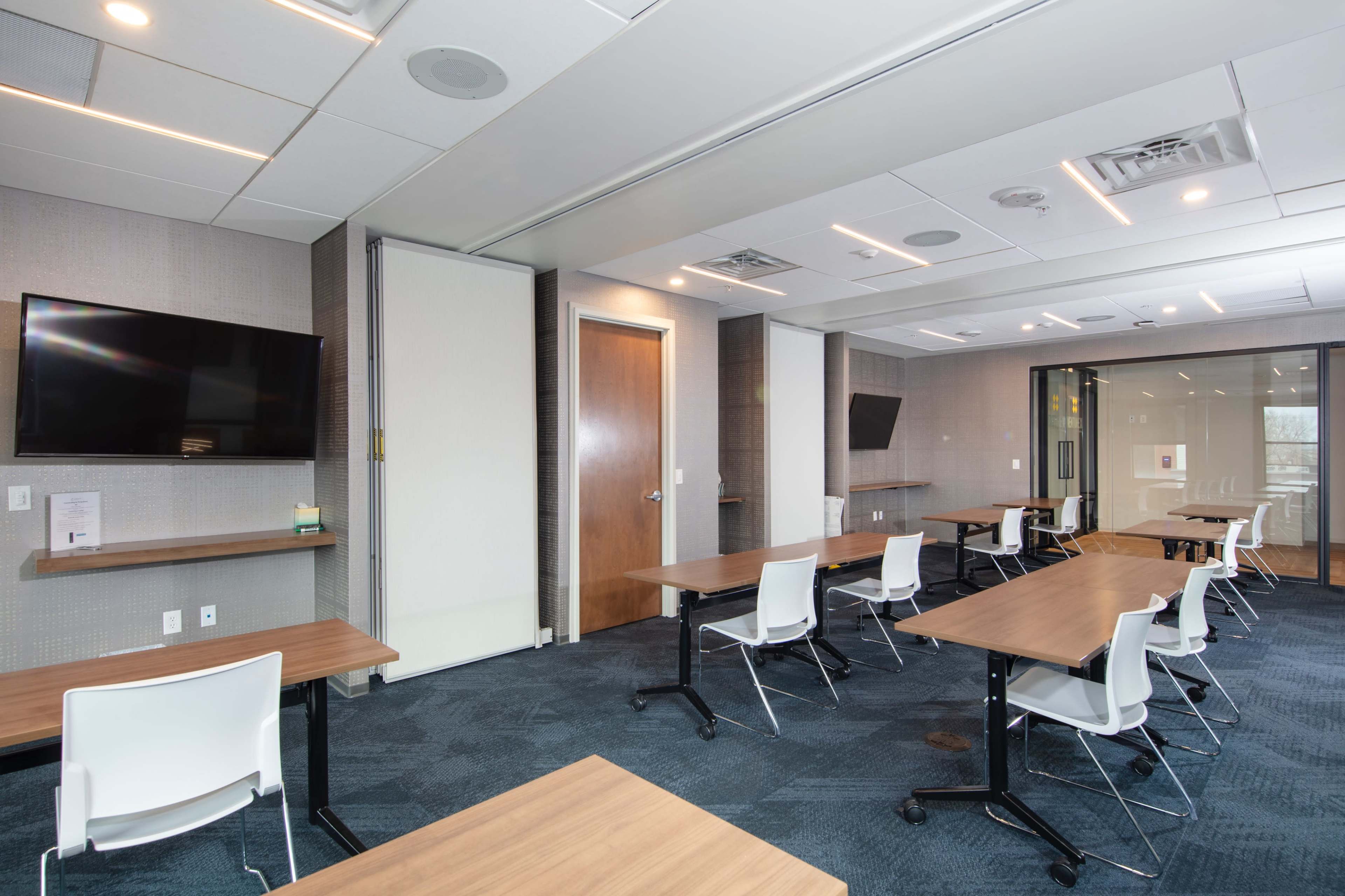 The image shows a modern conference room with wooden tables, white chairs, and two wall-mounted televisions.