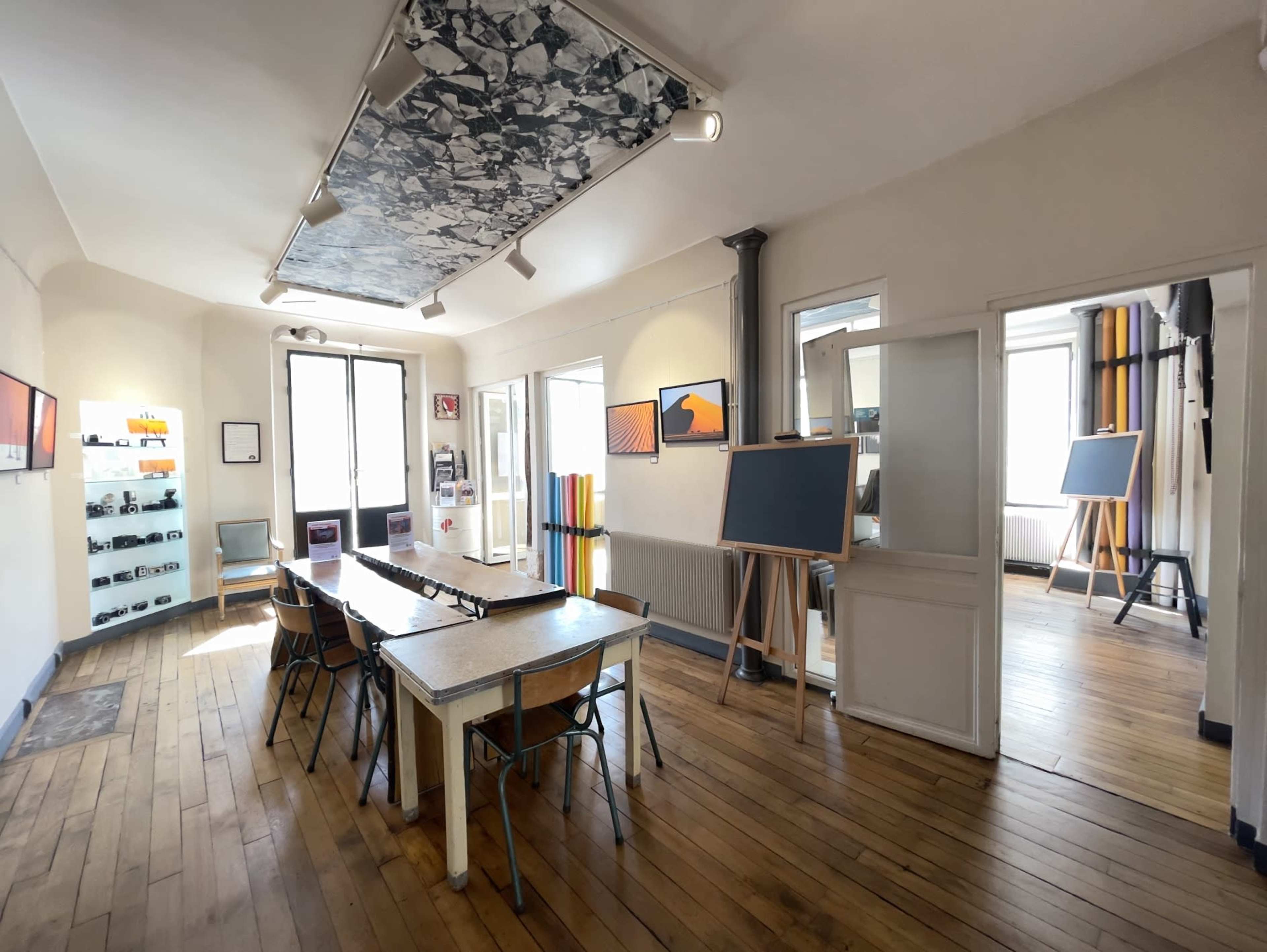A bright room with wooden floors, featuring a long table, several chairs, and easels with blank canvases against the walls.