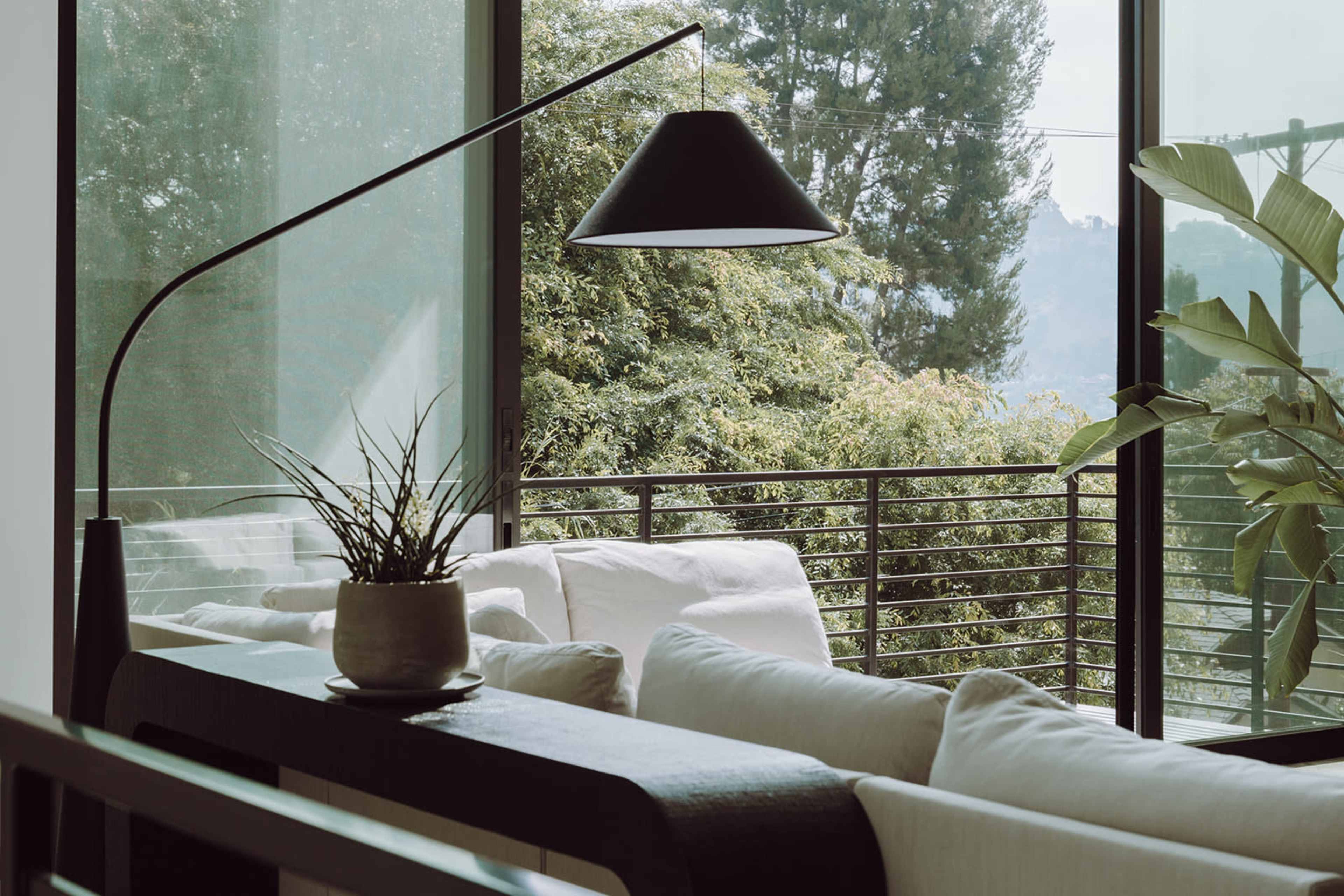 A modern living room features a sleek floor lamp, a potted plant, and a view of greenery through large windows.