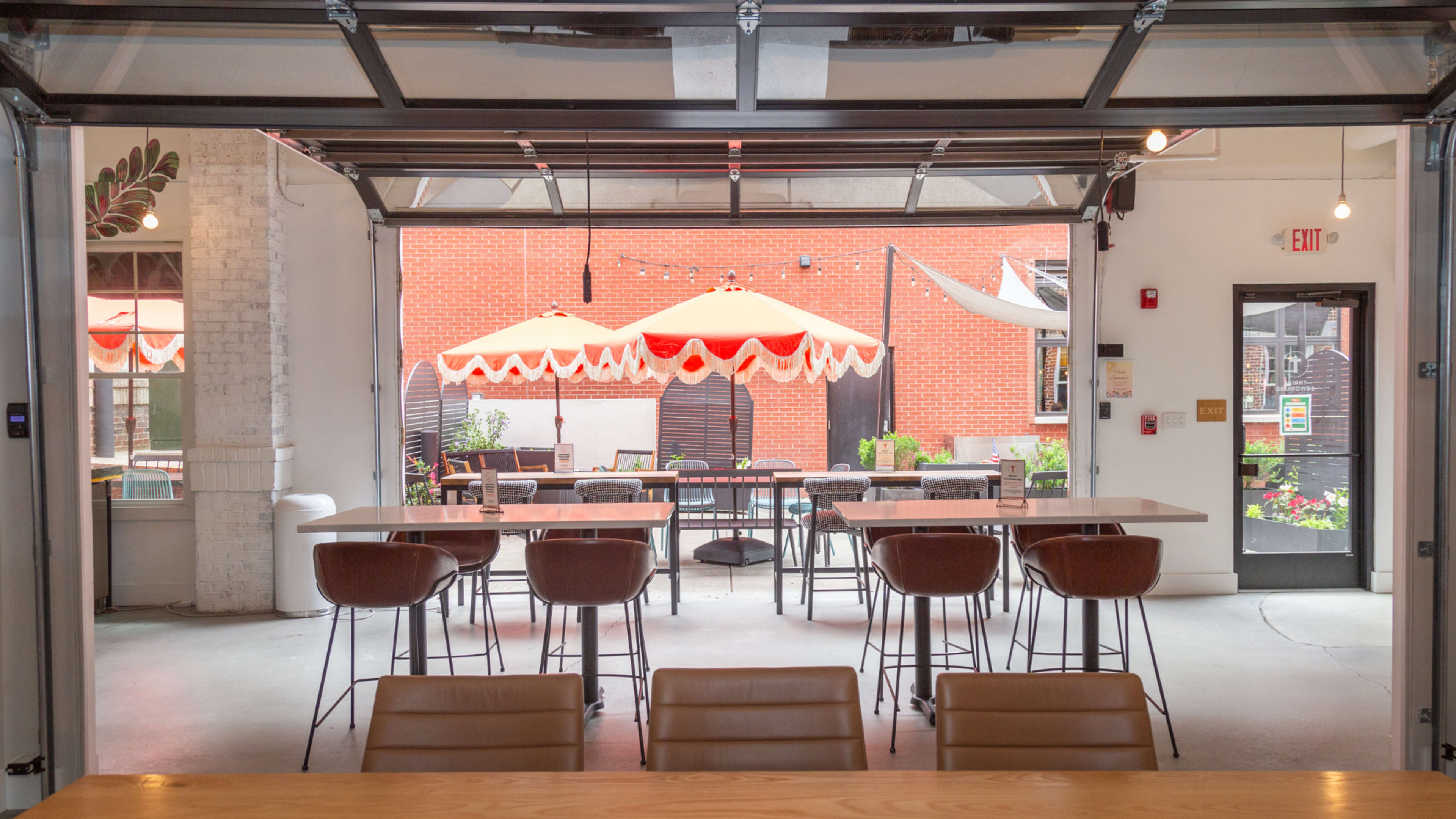 The image shows a modern indoor space with a large open door leading to an outdoor dining area equipped with umbrellas and tables.