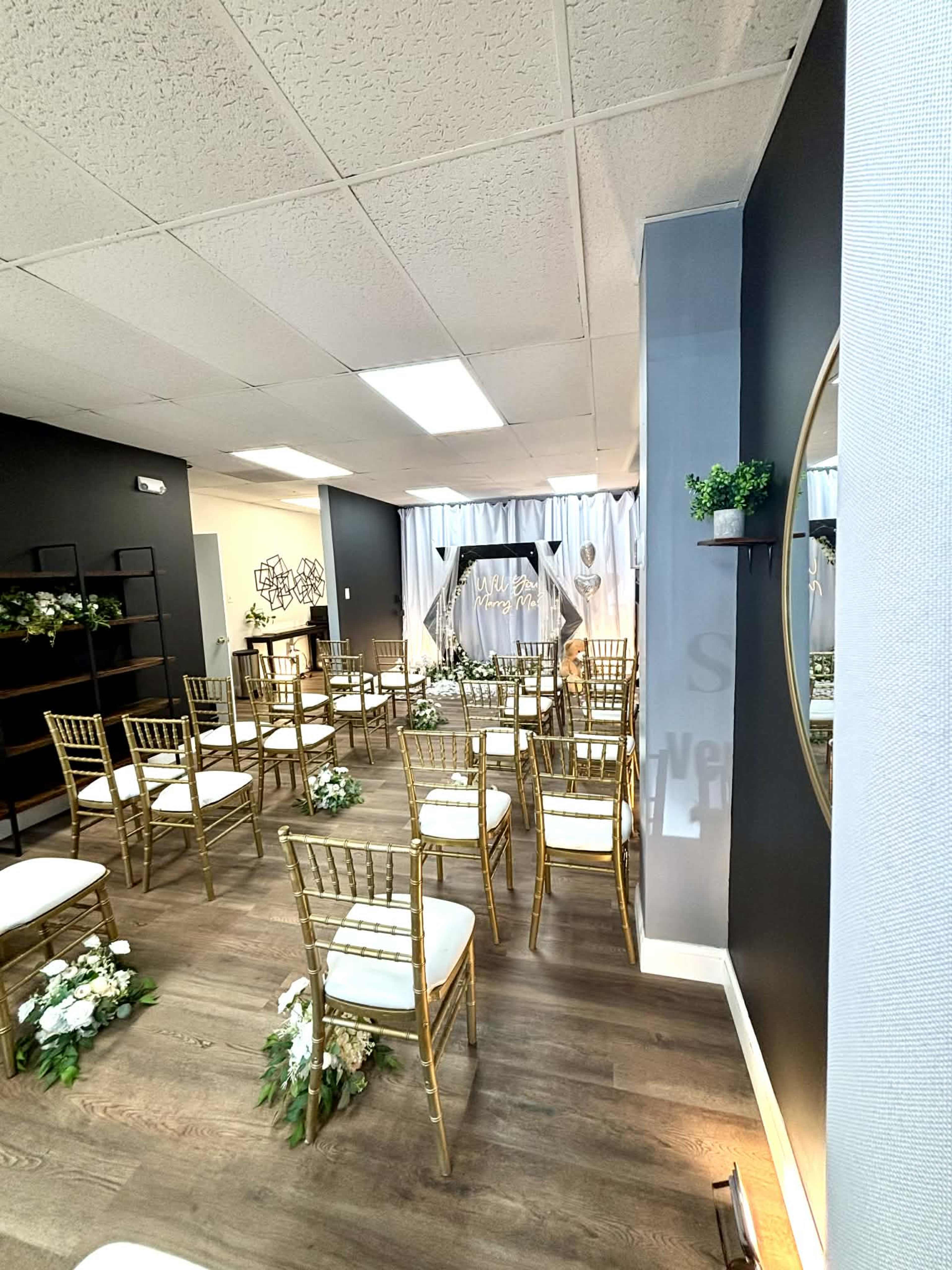 The image shows a decorated event space with rows of gold chiavari chairs, a floral arrangement, and a backdrop set up for a ceremony.