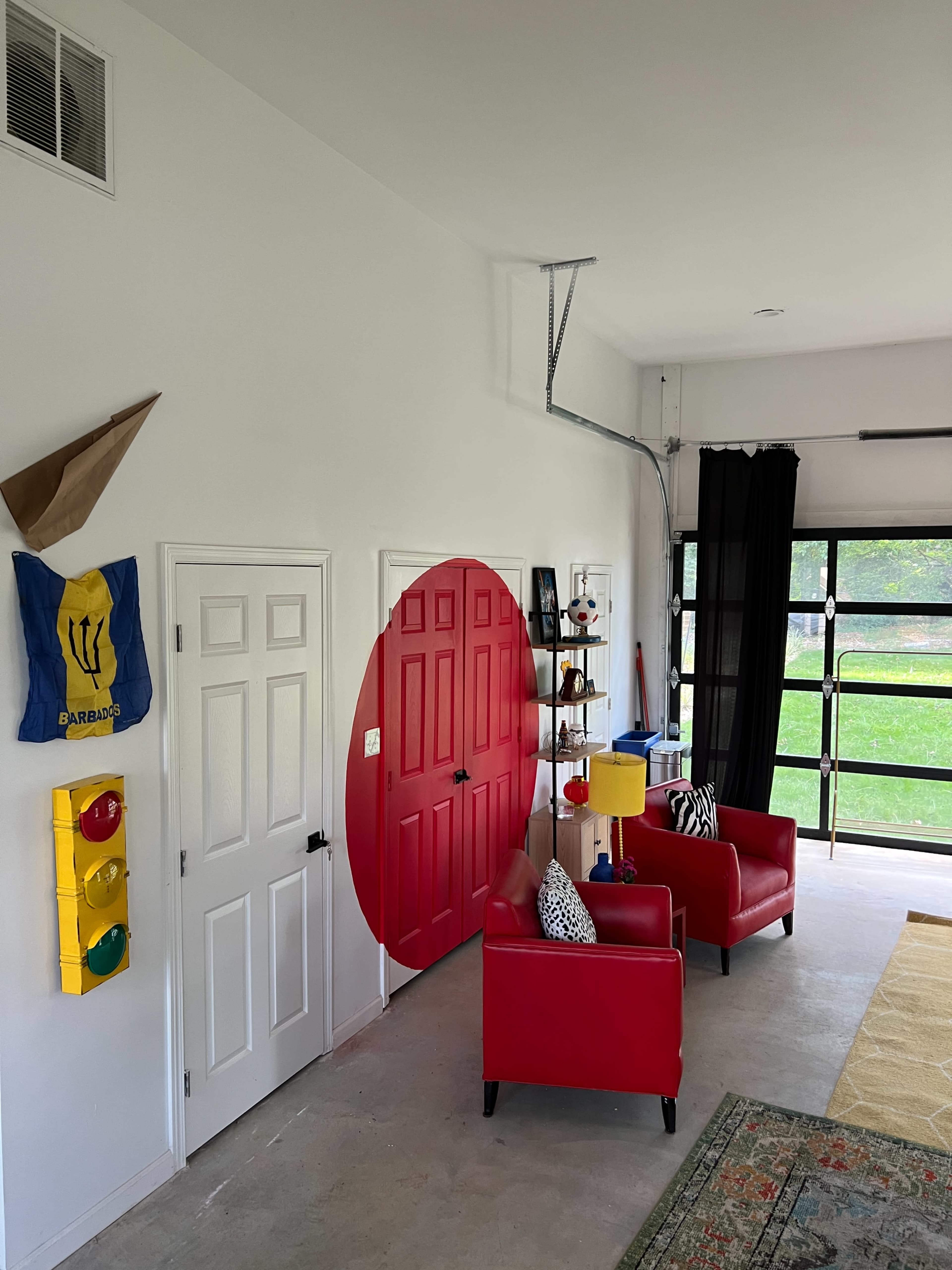 The image shows a modern garage space featuring red armchairs, a bright red door, a yellow lamp, and a decorative wall element resembling a large red circle.