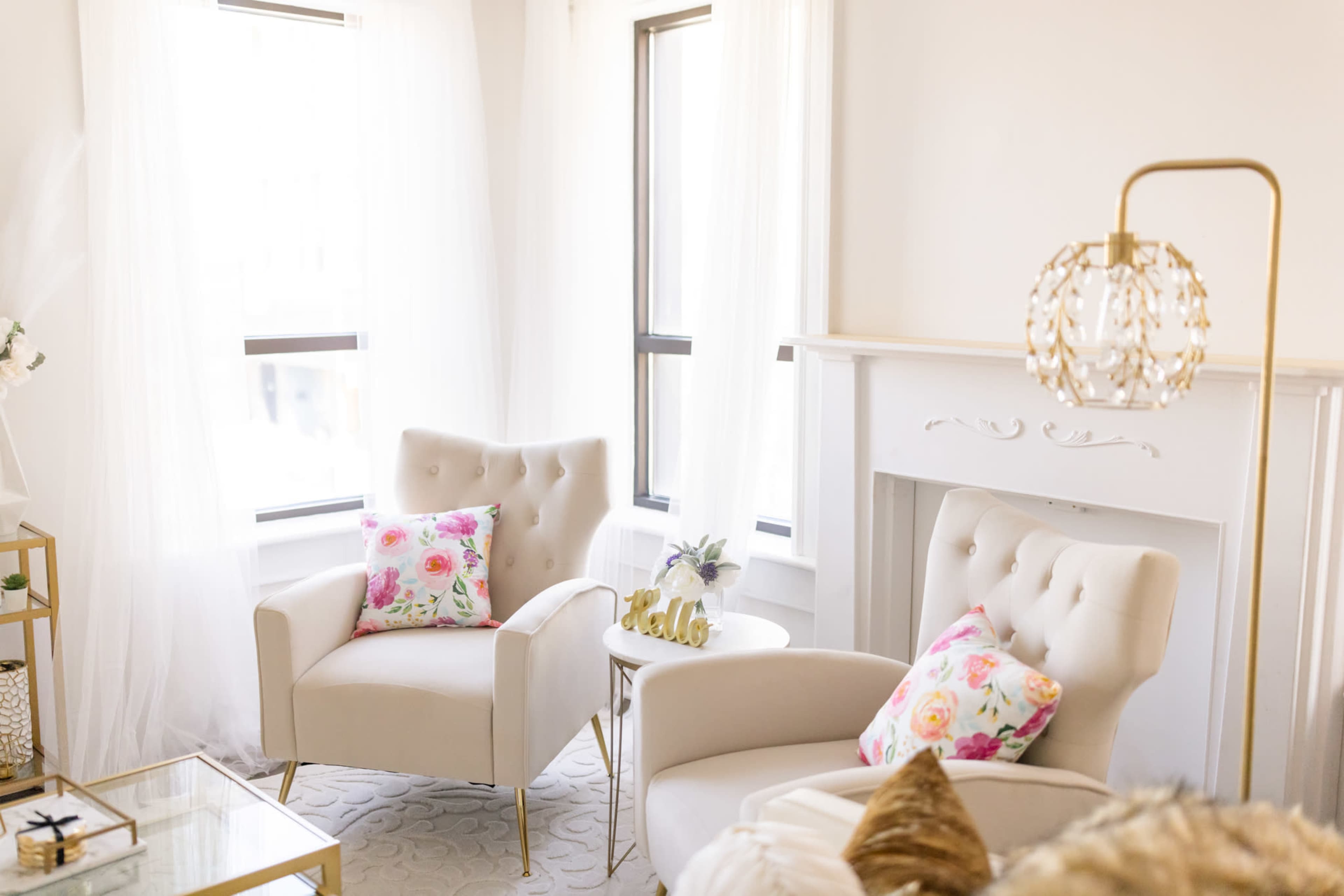 A bright room with two cream-colored chairs, both adorned with floral pillows, positioned beside a small round table and in front of a white fireplace.