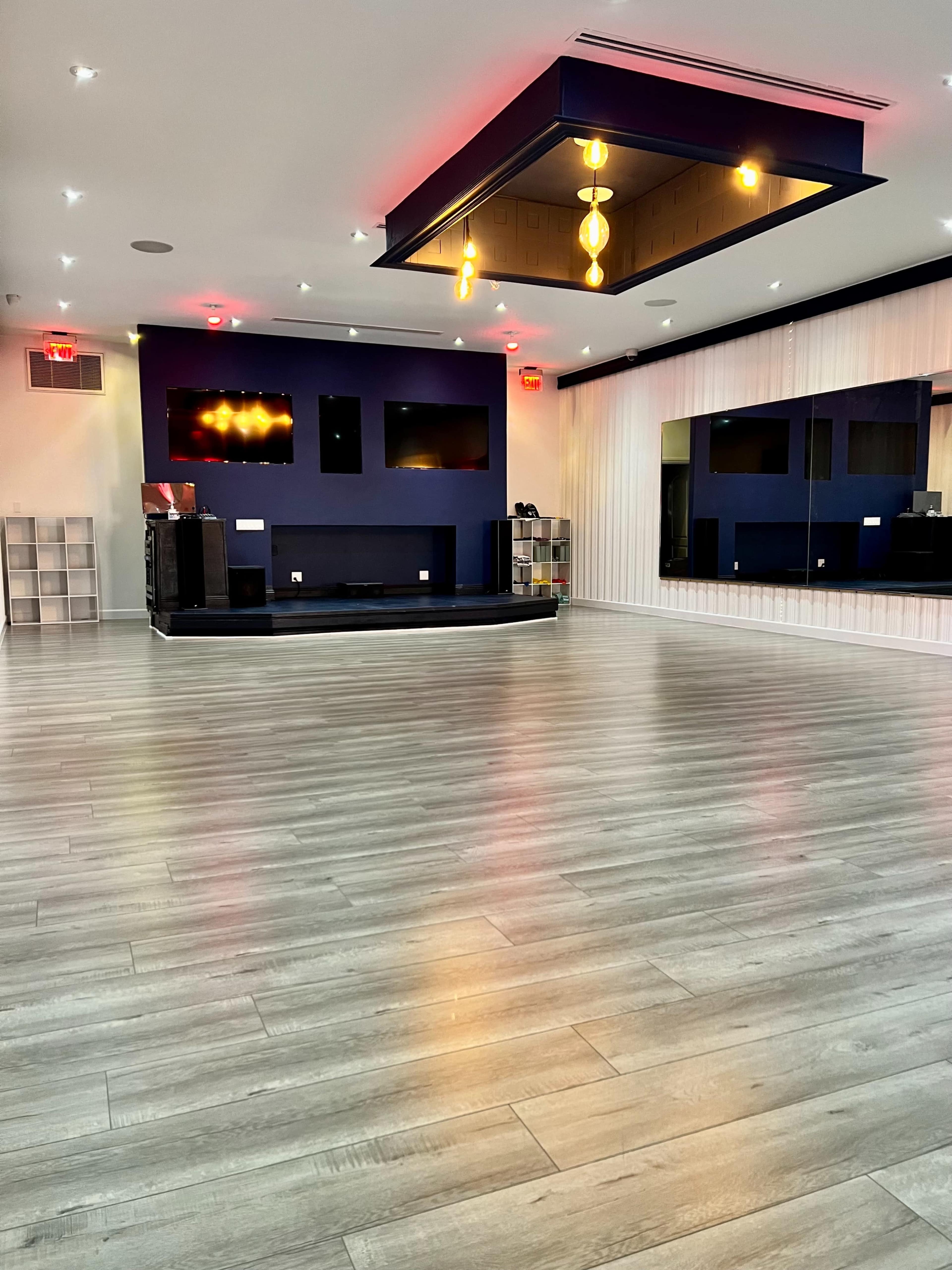 The image shows a spacious, modern interior with light wooden floors, a raised stage area, and large mirrors reflecting the walls painted in dark blue and white.