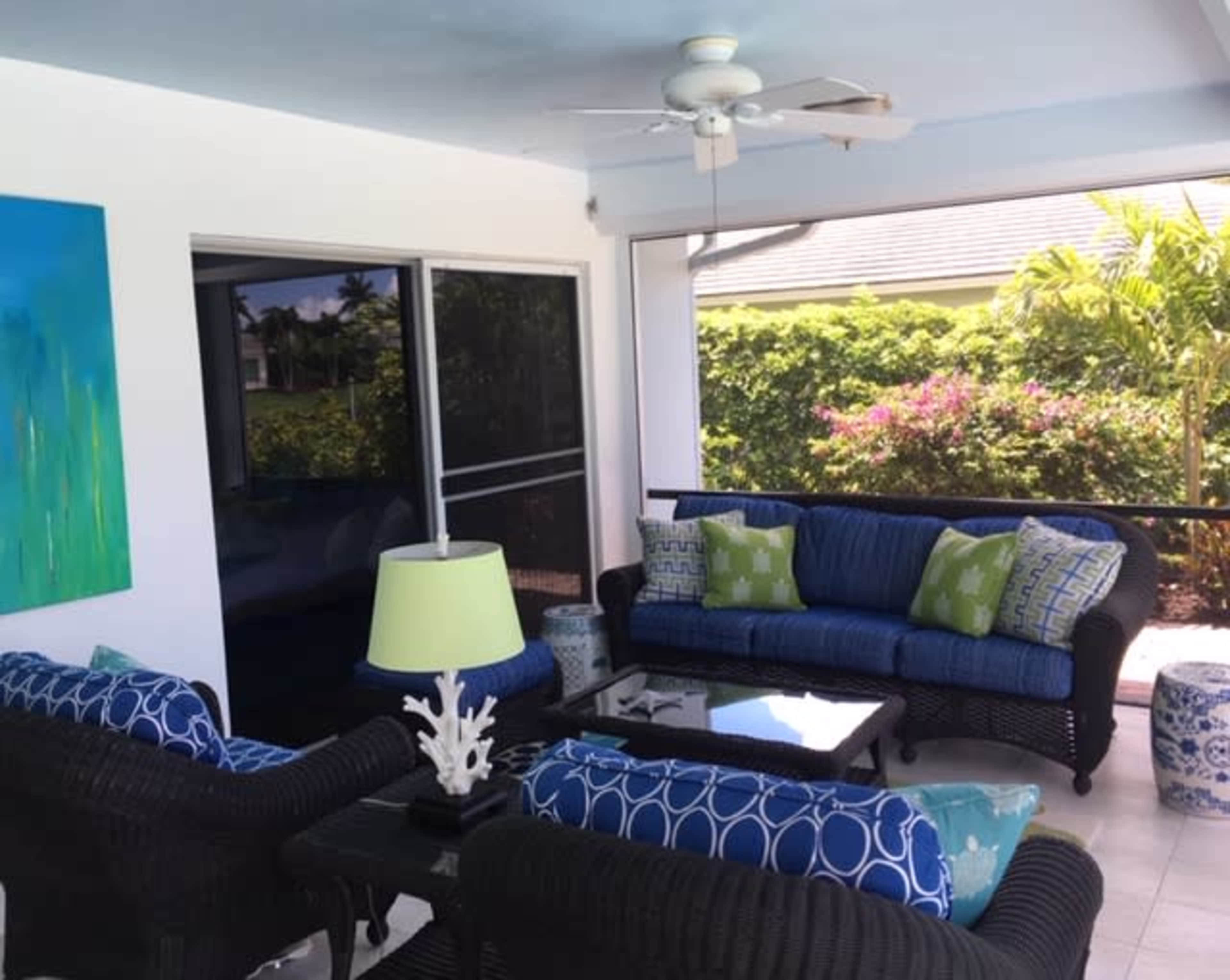 A bright, covered patio features wicker furniture with blue and green cushions, a glass table, and a lamp, surrounded by lush greenery.