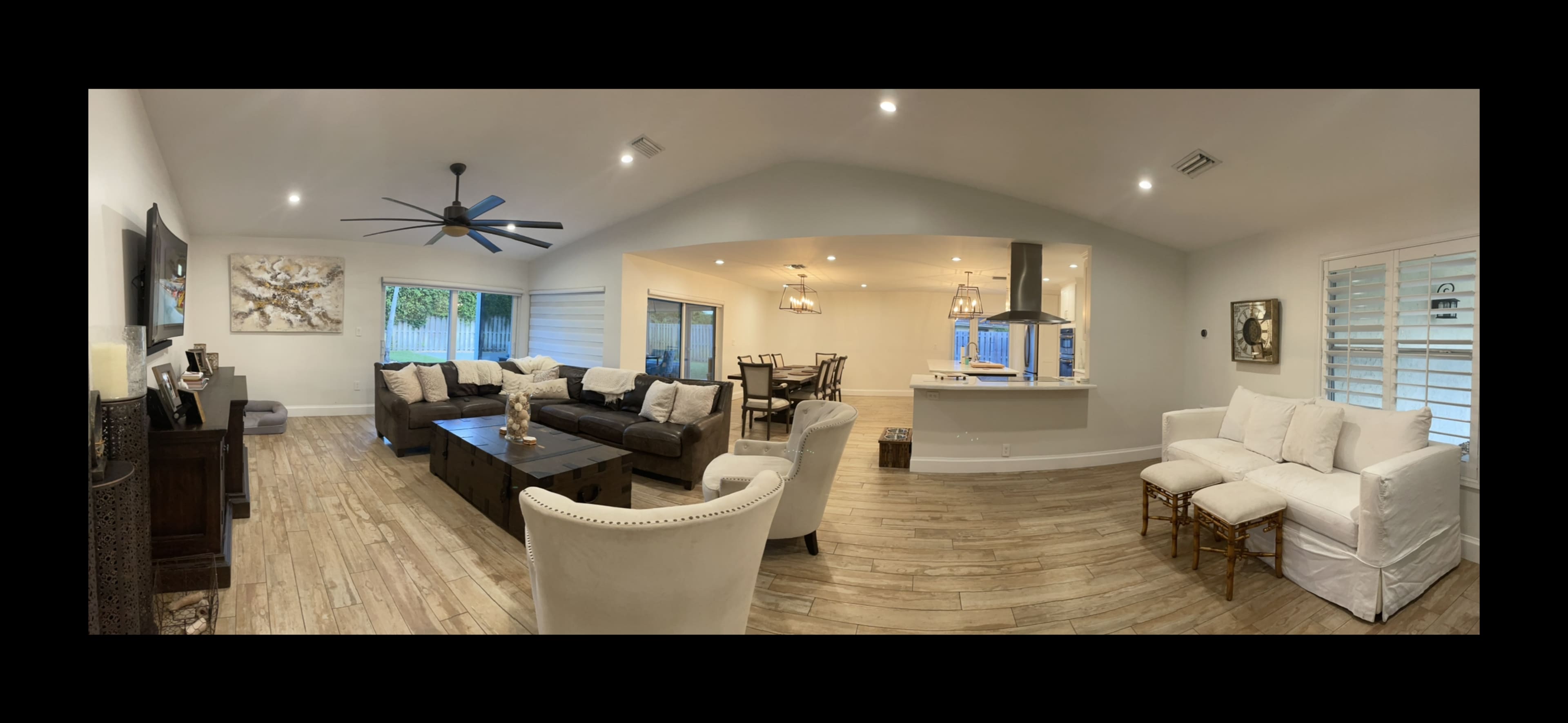 The image shows a spacious, modern living room and kitchen area with light-colored walls, a large sofa set, and a ceiling fan.
