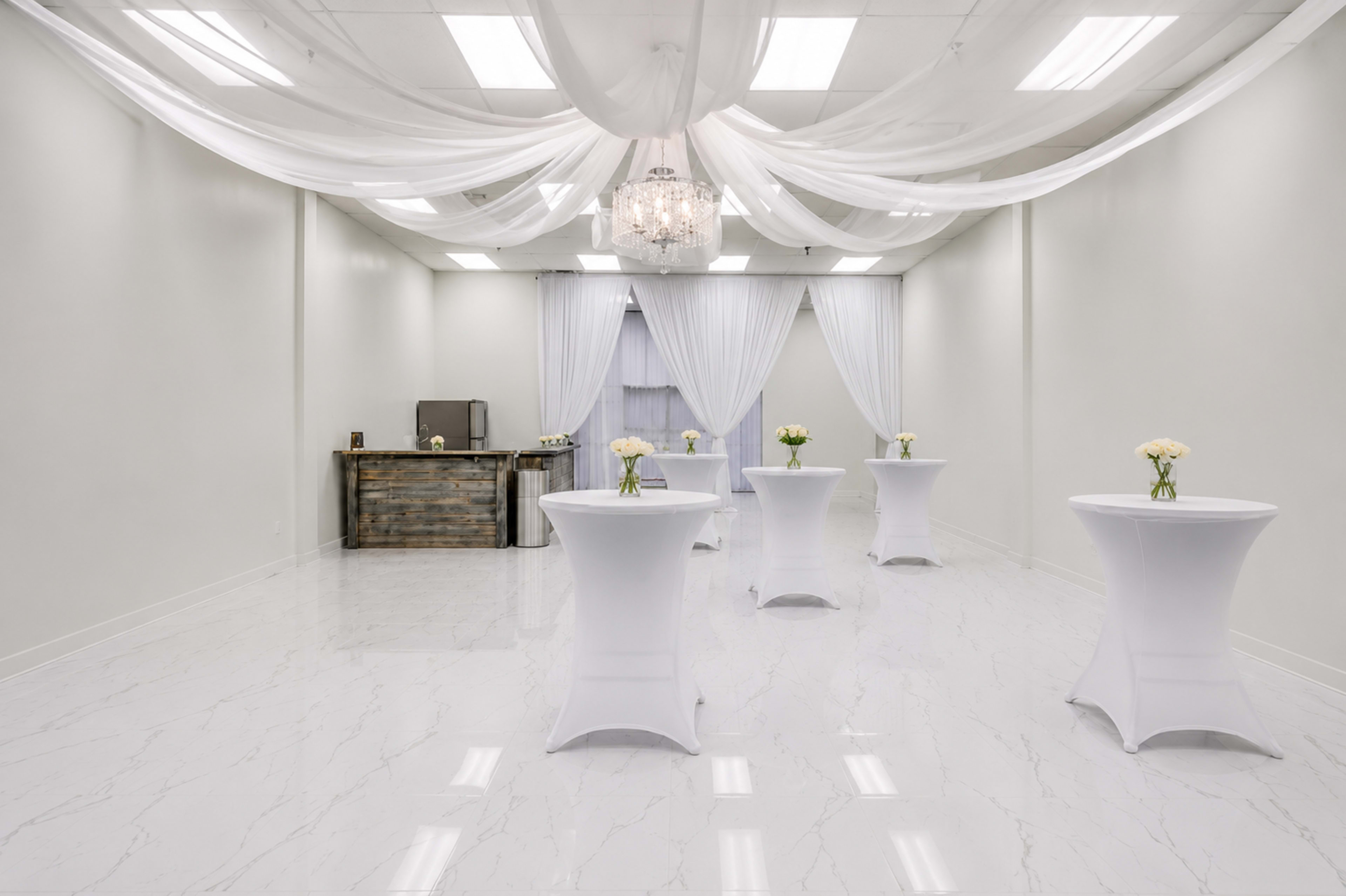 The image shows a spacious event room with a polished white floor, four round tables covered in white cloth, and a wooden bar set against a wall, all adorned with flower vases.
