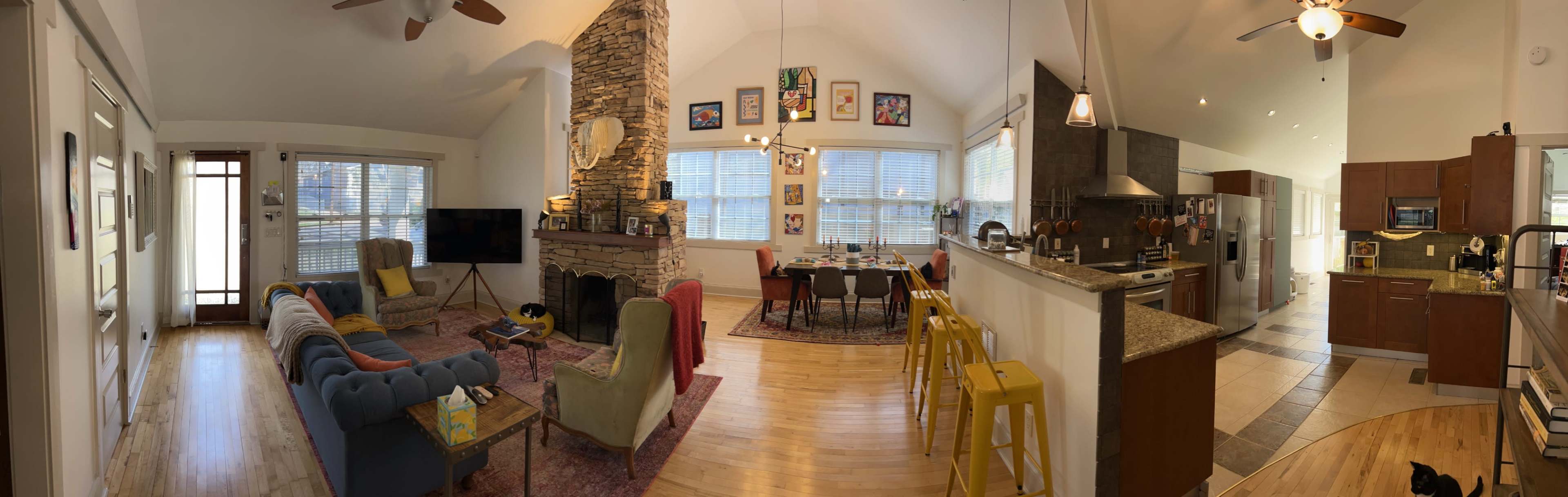 The image shows an open-concept living space featuring a living area with colorful chairs, a stone fireplace, a dining table, and a kitchen with wooden cabinets.