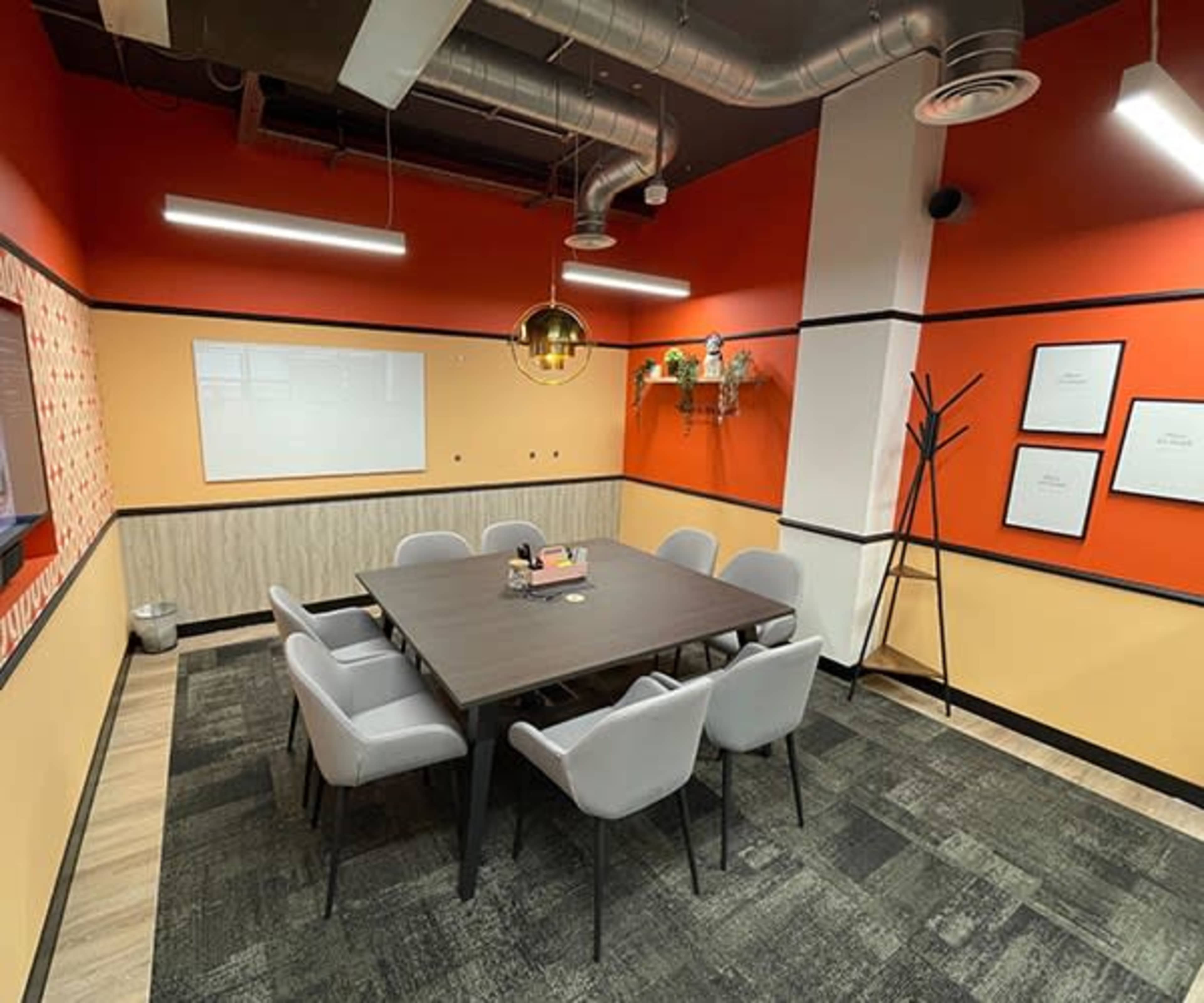 A modern meeting room with a large table surrounded by eight chairs, accented by orange and beige walls and decorative plants.