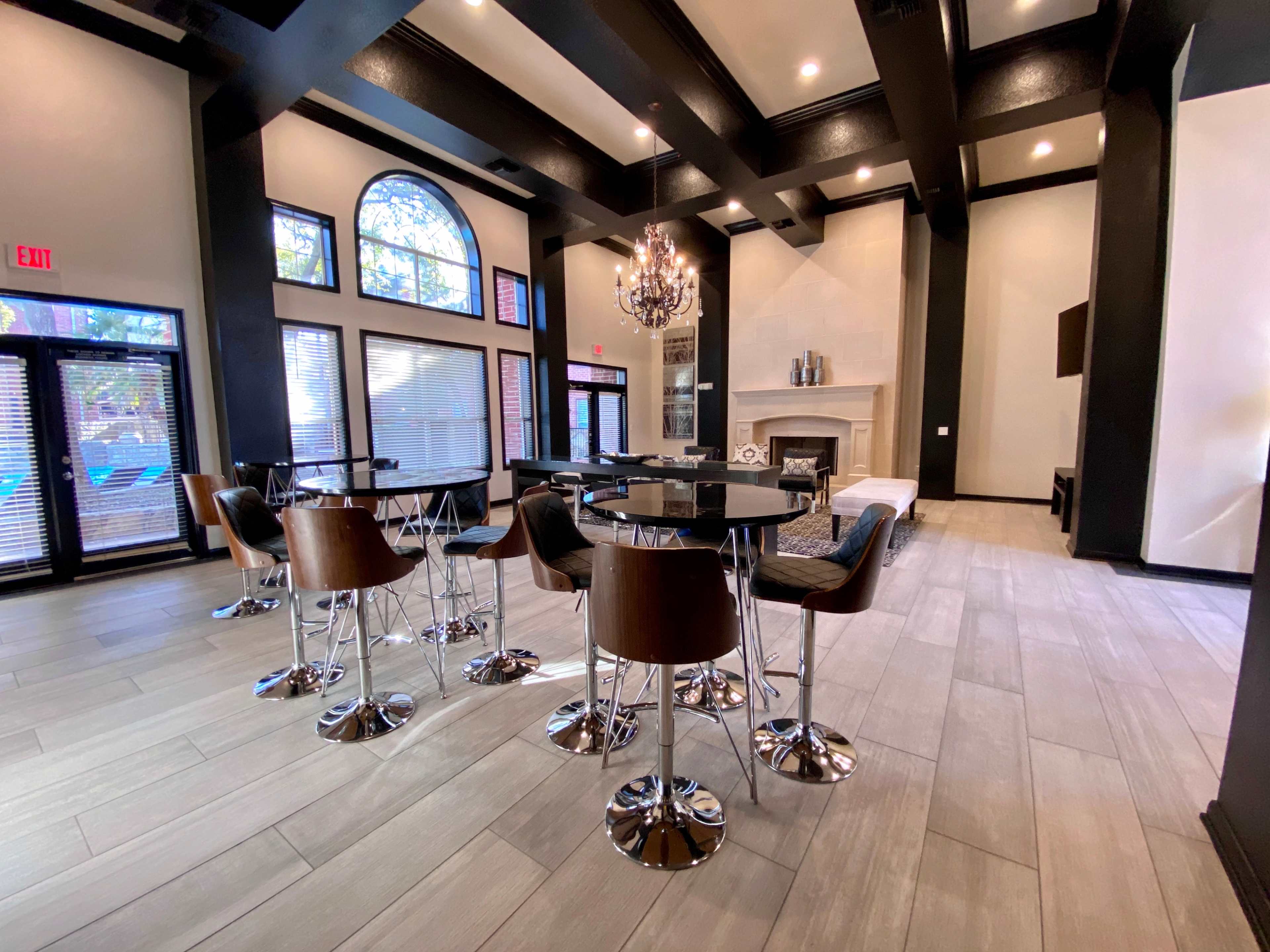 The image shows a modern, spacious lounge area with high ceilings, large windows, and several round tables surrounded by chairs.