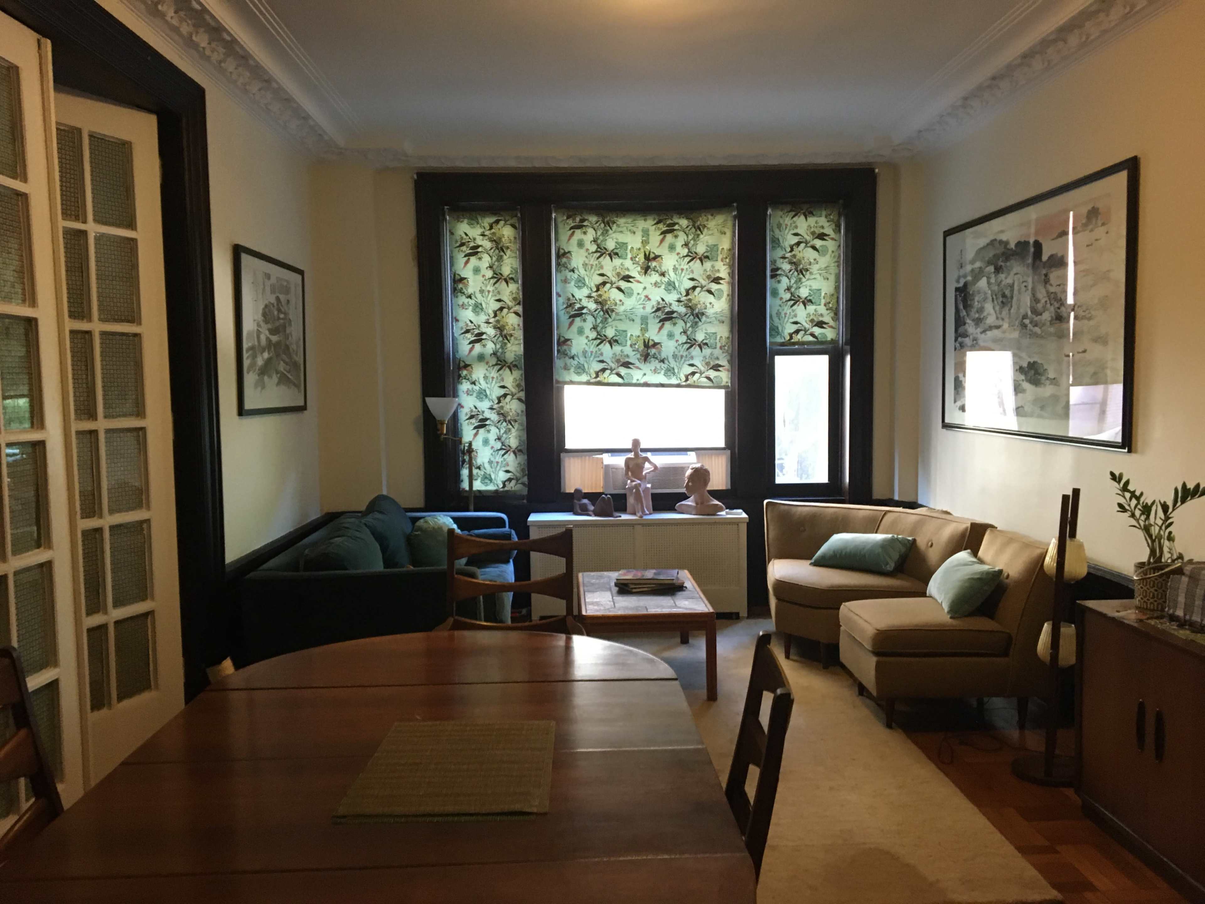A living room features a mix of seating, a dining table, and large windows with patterned curtains.