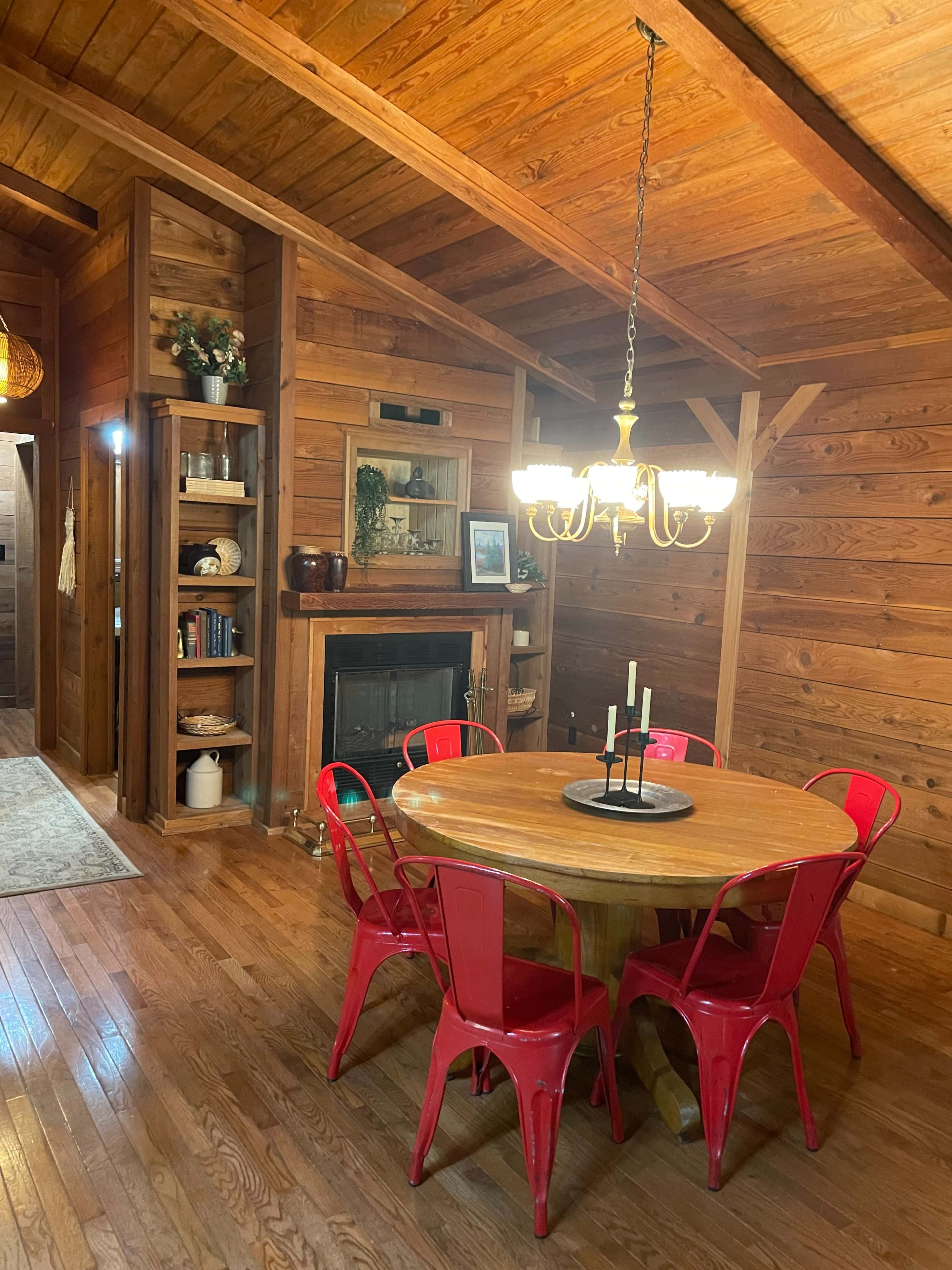 The image shows a wooden dining area featuring a round table surrounded by red chairs, with a chandelier overhead and a fireplace lined with decorative items.