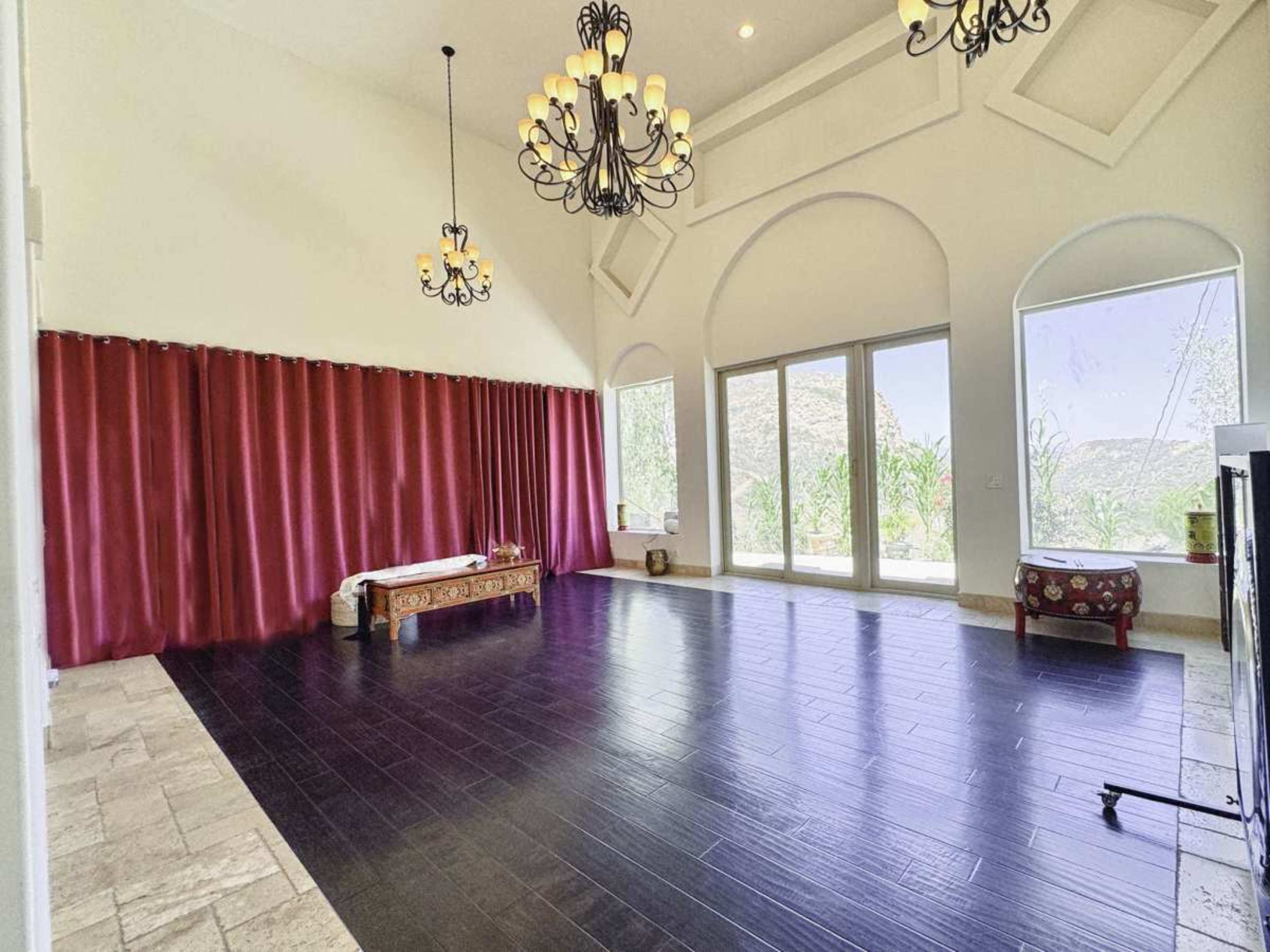 The image shows a spacious room with high ceilings, large windows, and a combination of dark hardwood and stone flooring, accented by red curtains and decorative lighting fixtures.