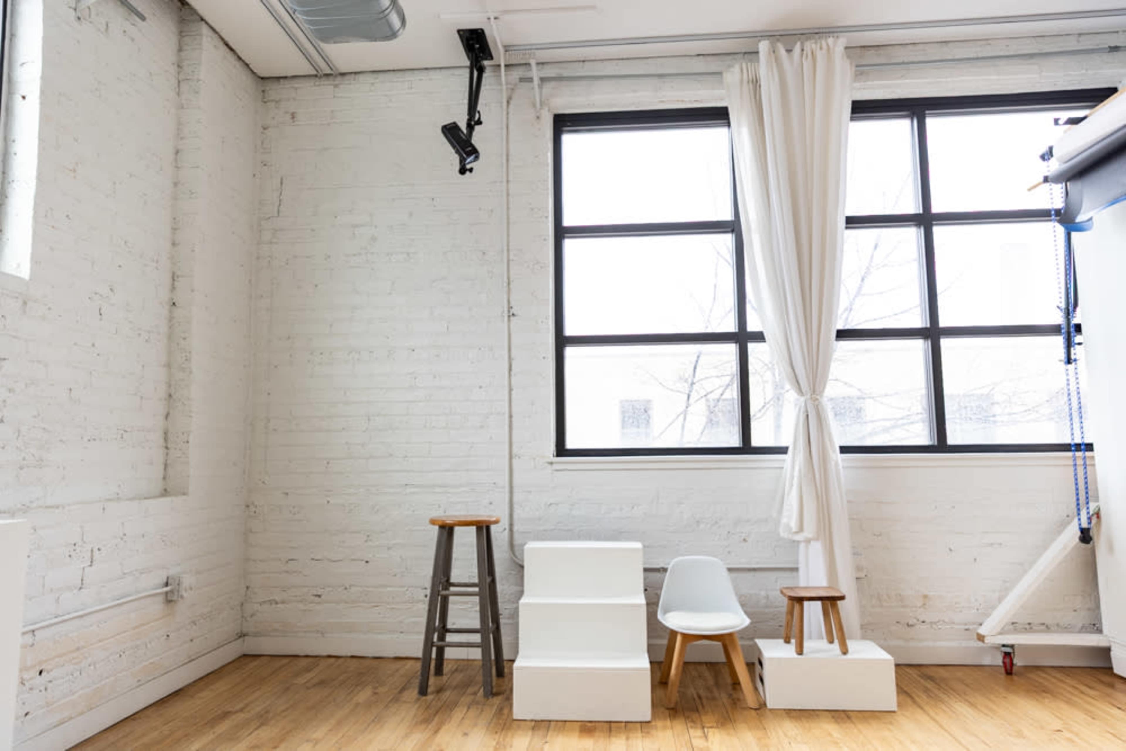 The image shows a minimalist interior space with white brick walls, large windows, and a few simple wooden stools along with a white chair.