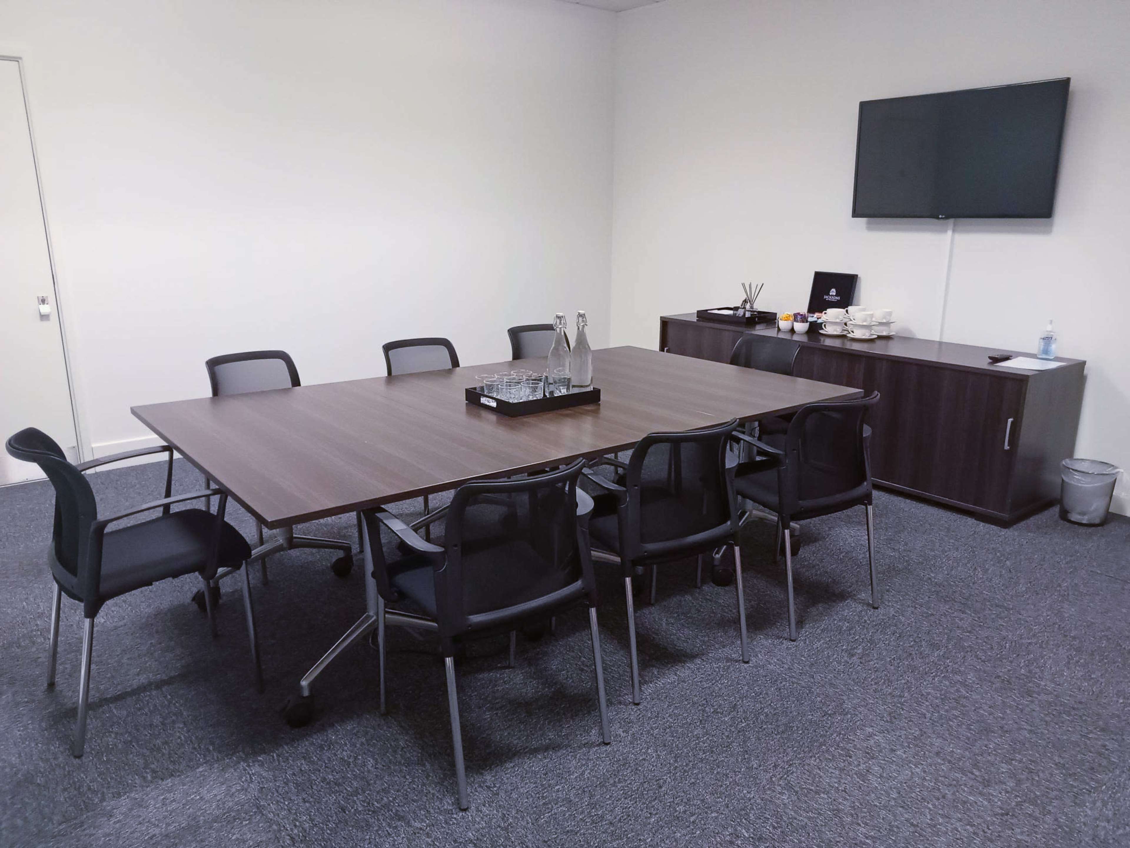 A conference room features a large rectangular table surrounded by eight chairs, with a television mounted on the wall and refreshments on a side cabinet.