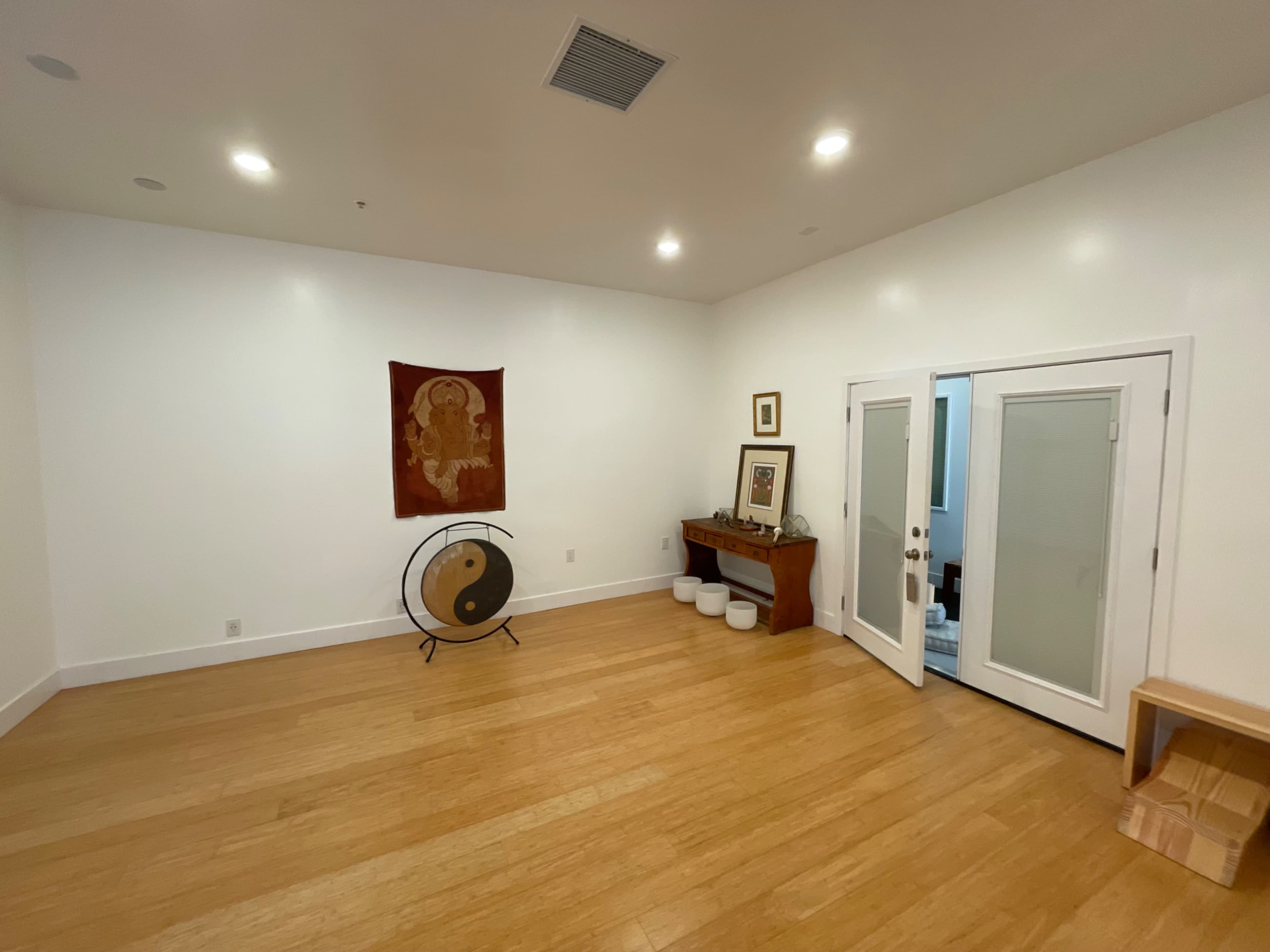 A spacious room with wooden flooring, decorated with a hanging tapestry, a gong on a stand, and a wooden console table near a set of glass-panel doors.