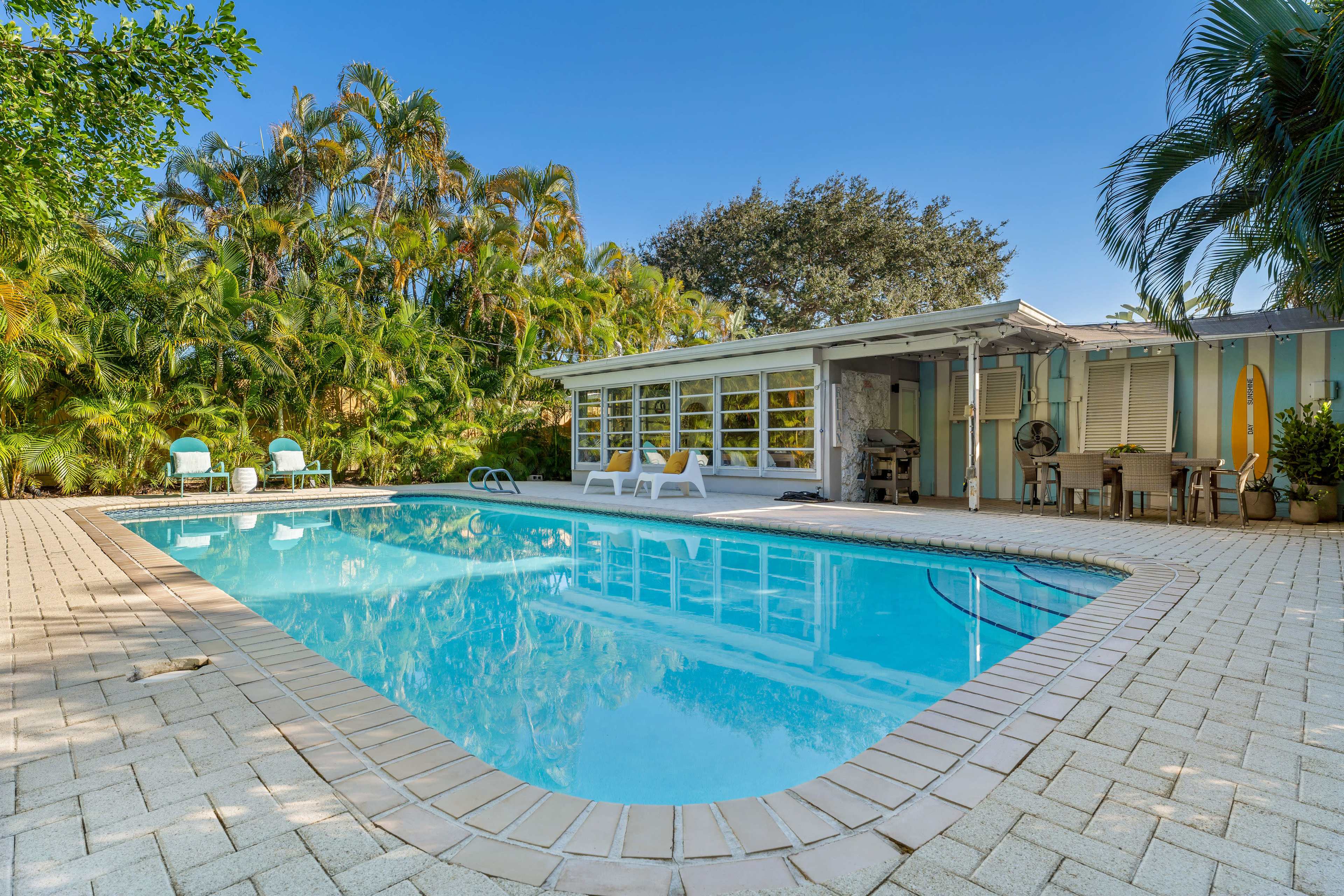 A clear blue swimming pool is surrounded by patio tiles and palm trees, with a house featuring large windows in the background.