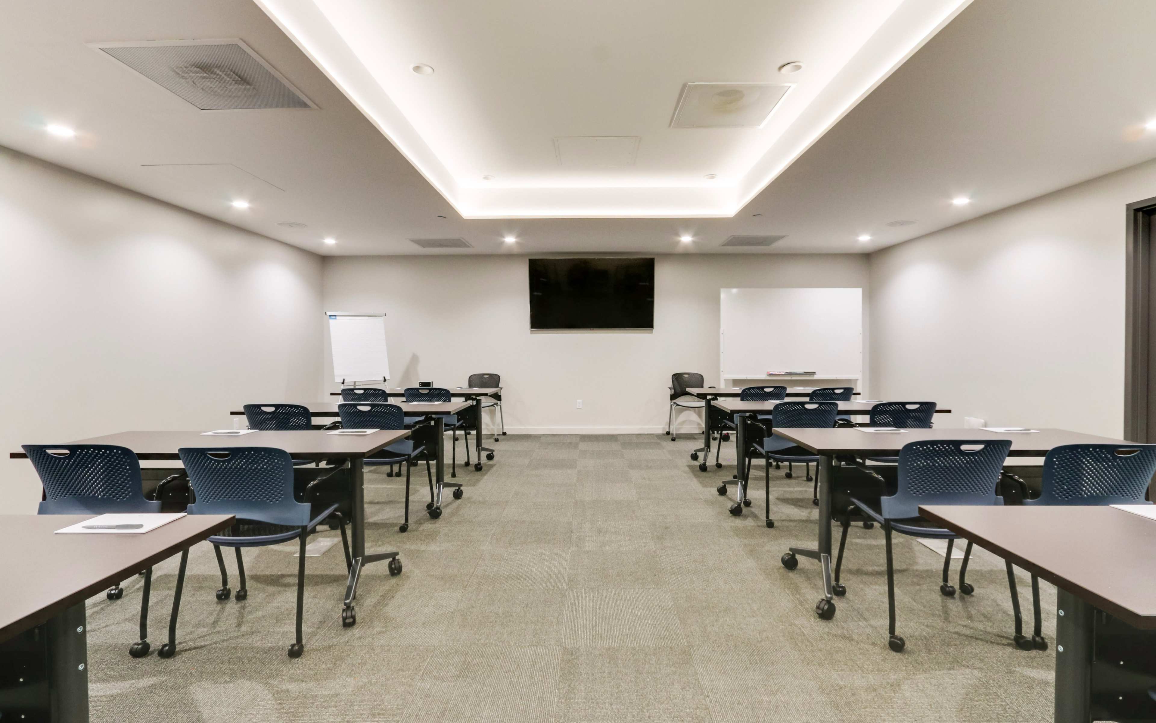 A modern conference room featuring several rows of tables and chairs, a television screen on the wall, and a whiteboard.