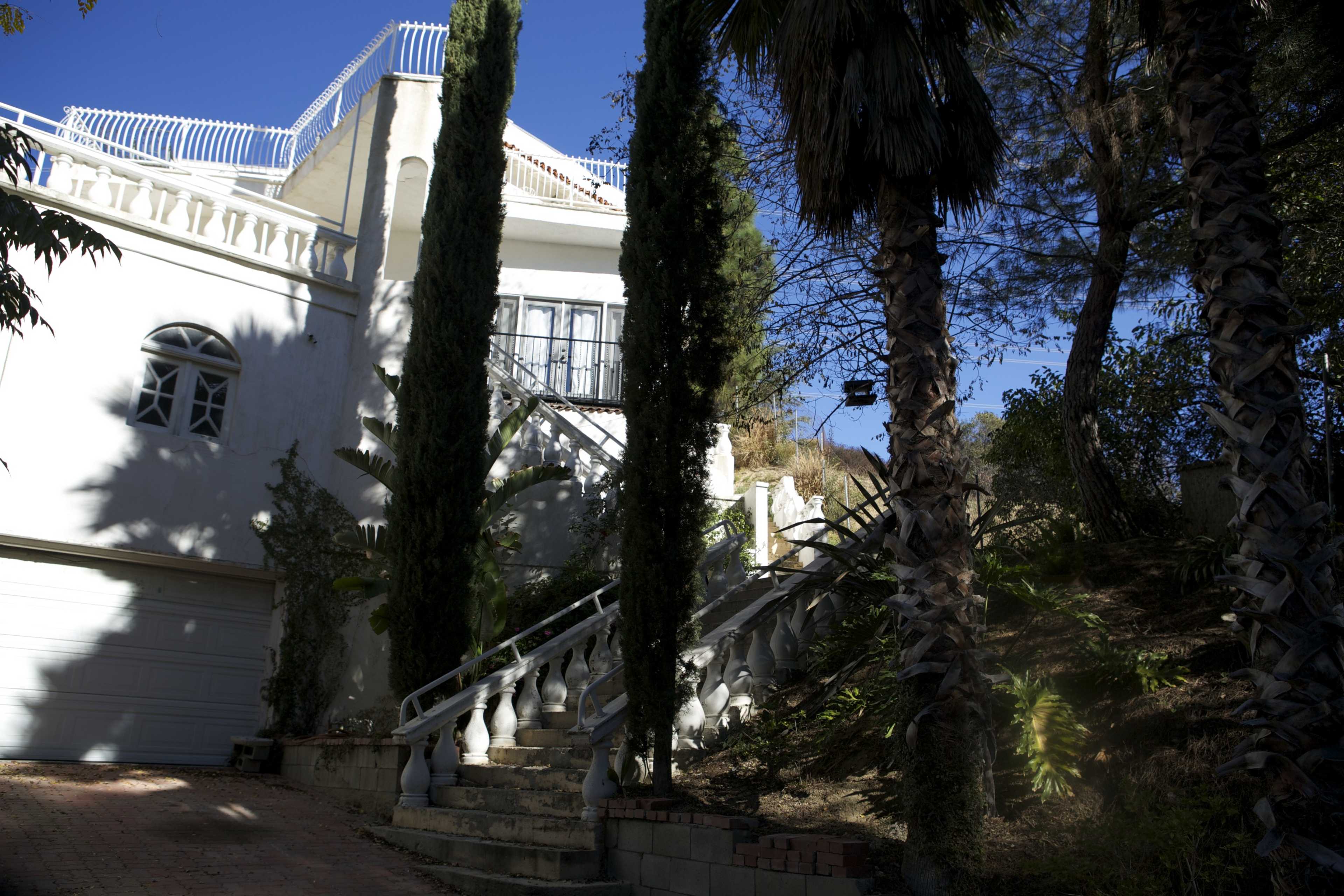 A white house with a balcony and stairs flanked by palm trees, set against a clear blue sky.