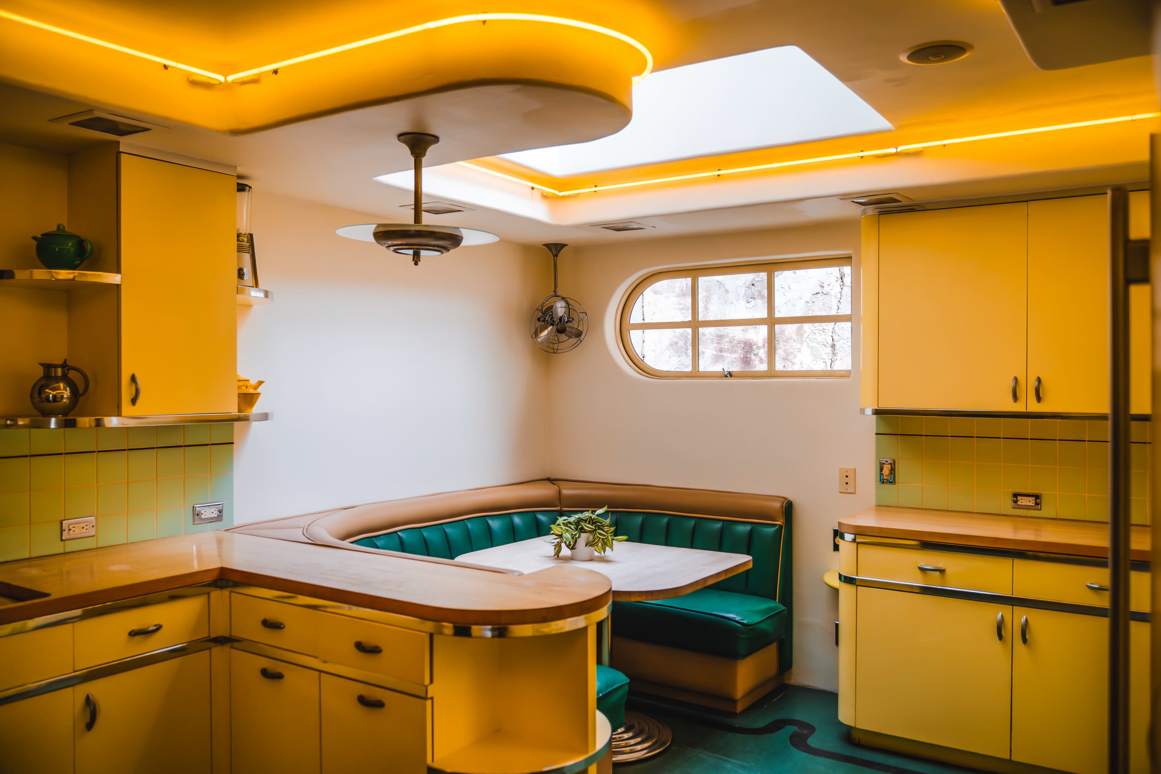 The image shows a retro-style kitchen featuring yellow cabinetry, a green upholstered booth, and a skylight.