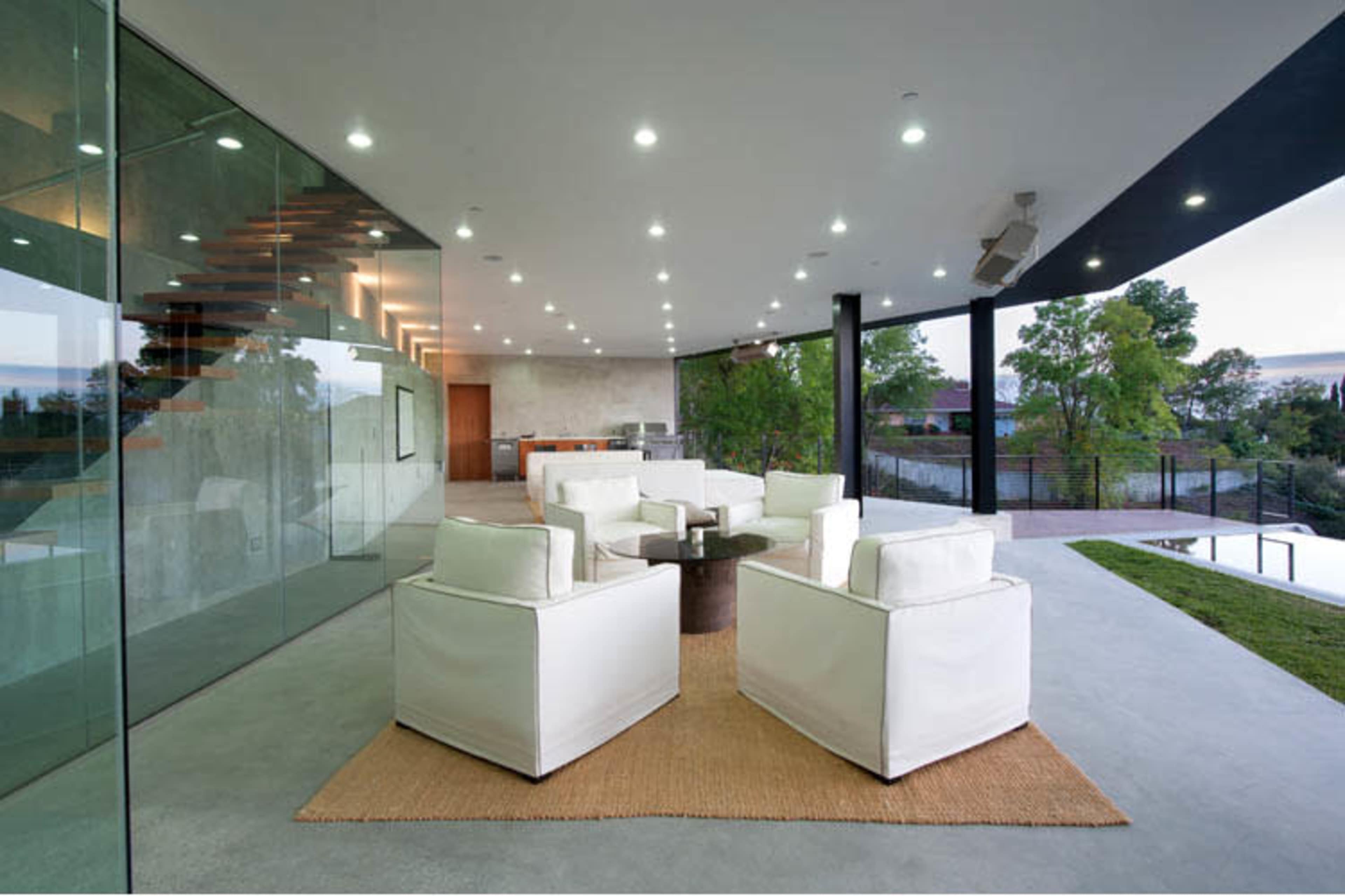 The image shows a modern, open-area living space with white furniture arranged on a woven rug and large glass windows revealing a view of greenery outside.