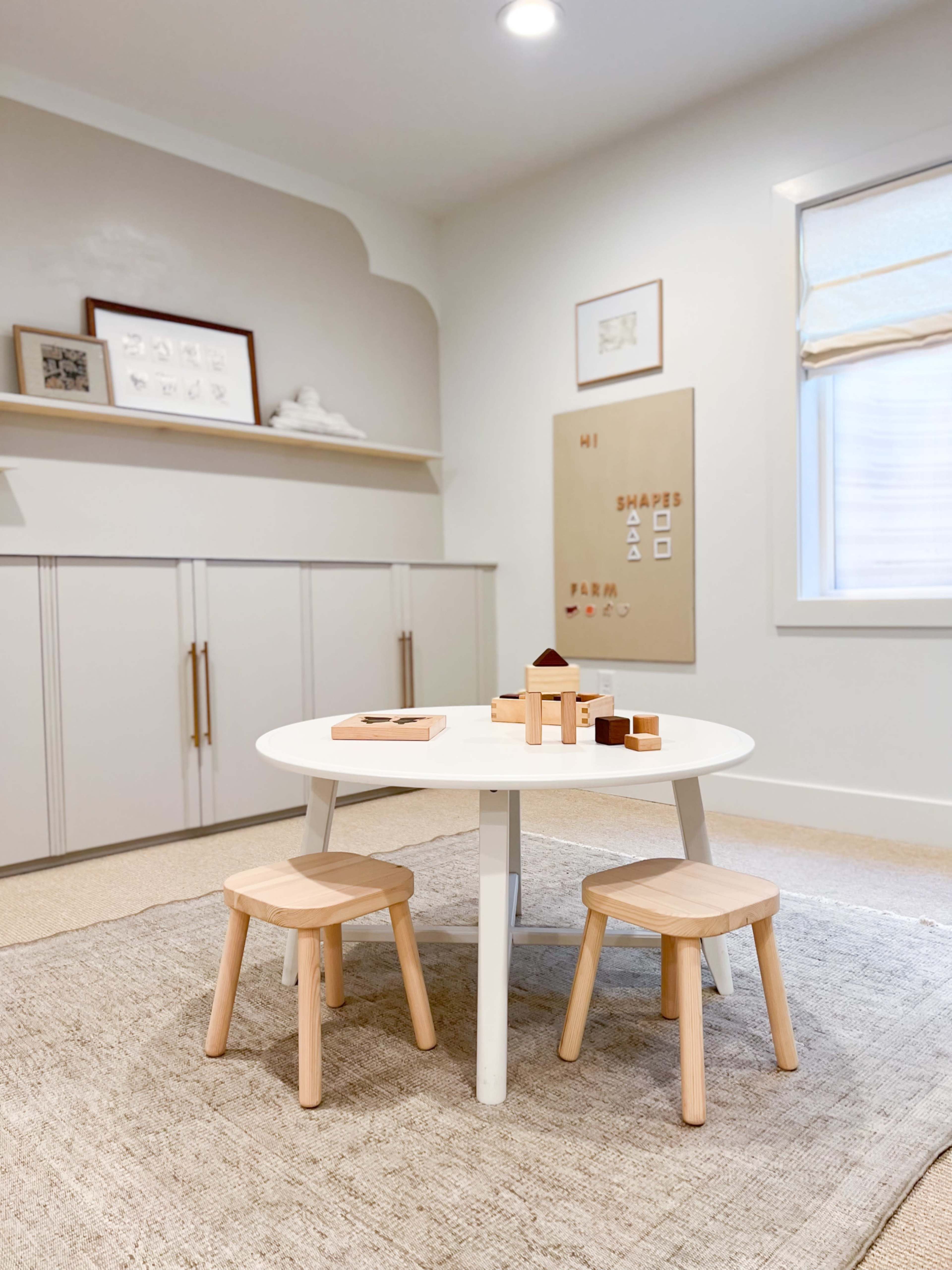 A small, round white table with two wooden stools is set in a minimalist room featuring light-colored walls and shelves with decorative items.
