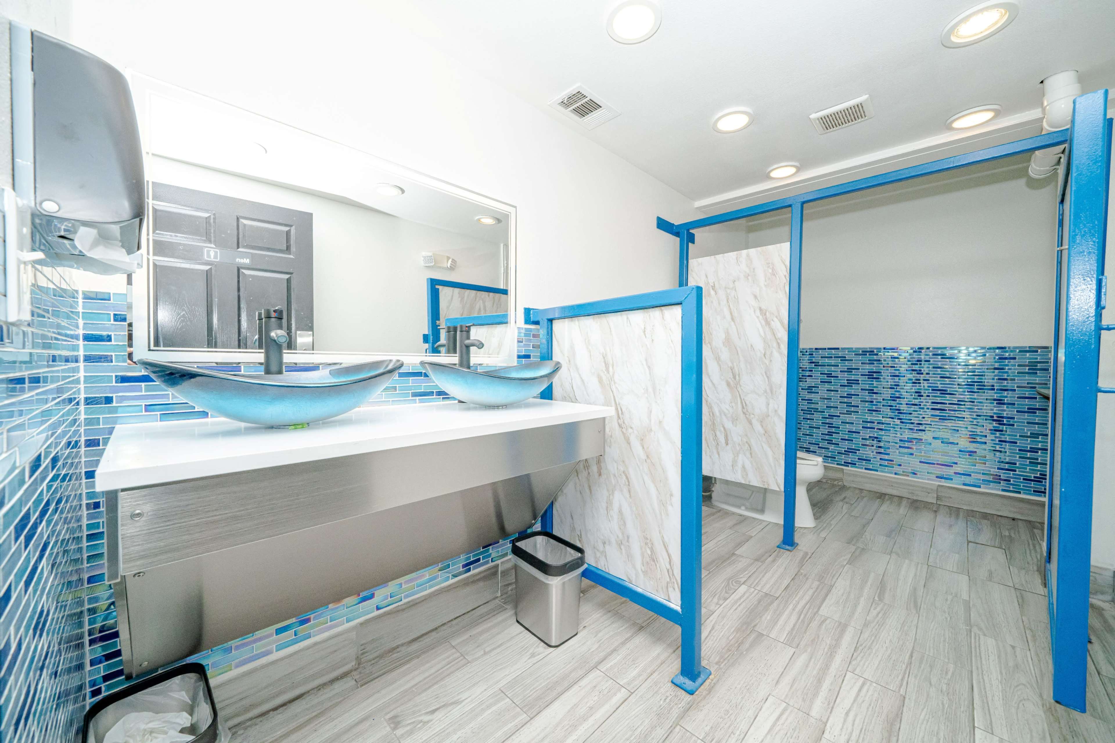 The image shows a modern bathroom with a large mirror, two blue vessel sinks, a partitioned toilet area, and tiled walls featuring a blue mosaic design.