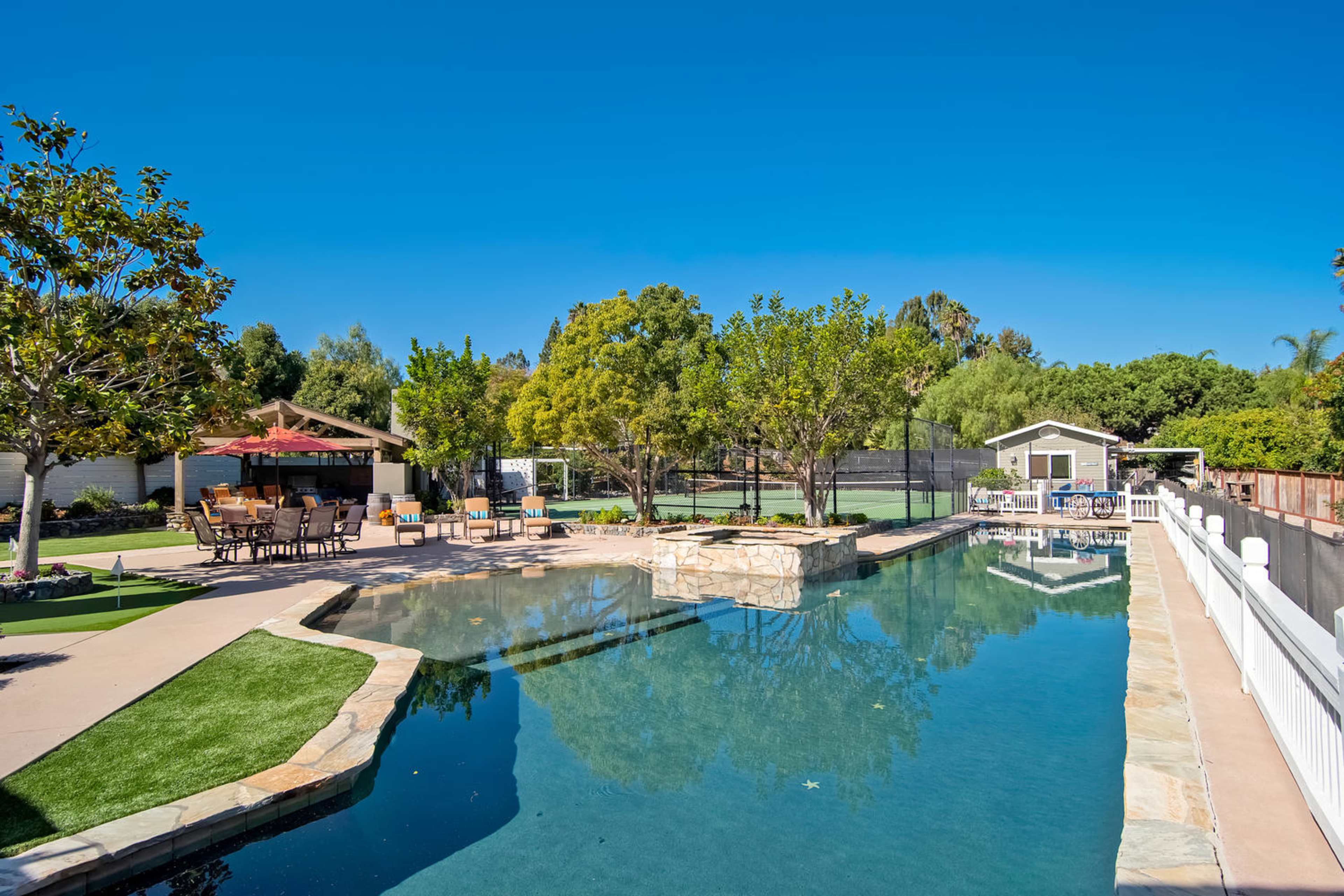 The image features a swimming pool with stone accents, surrounded by landscaped greenery and outdoor seating areas.