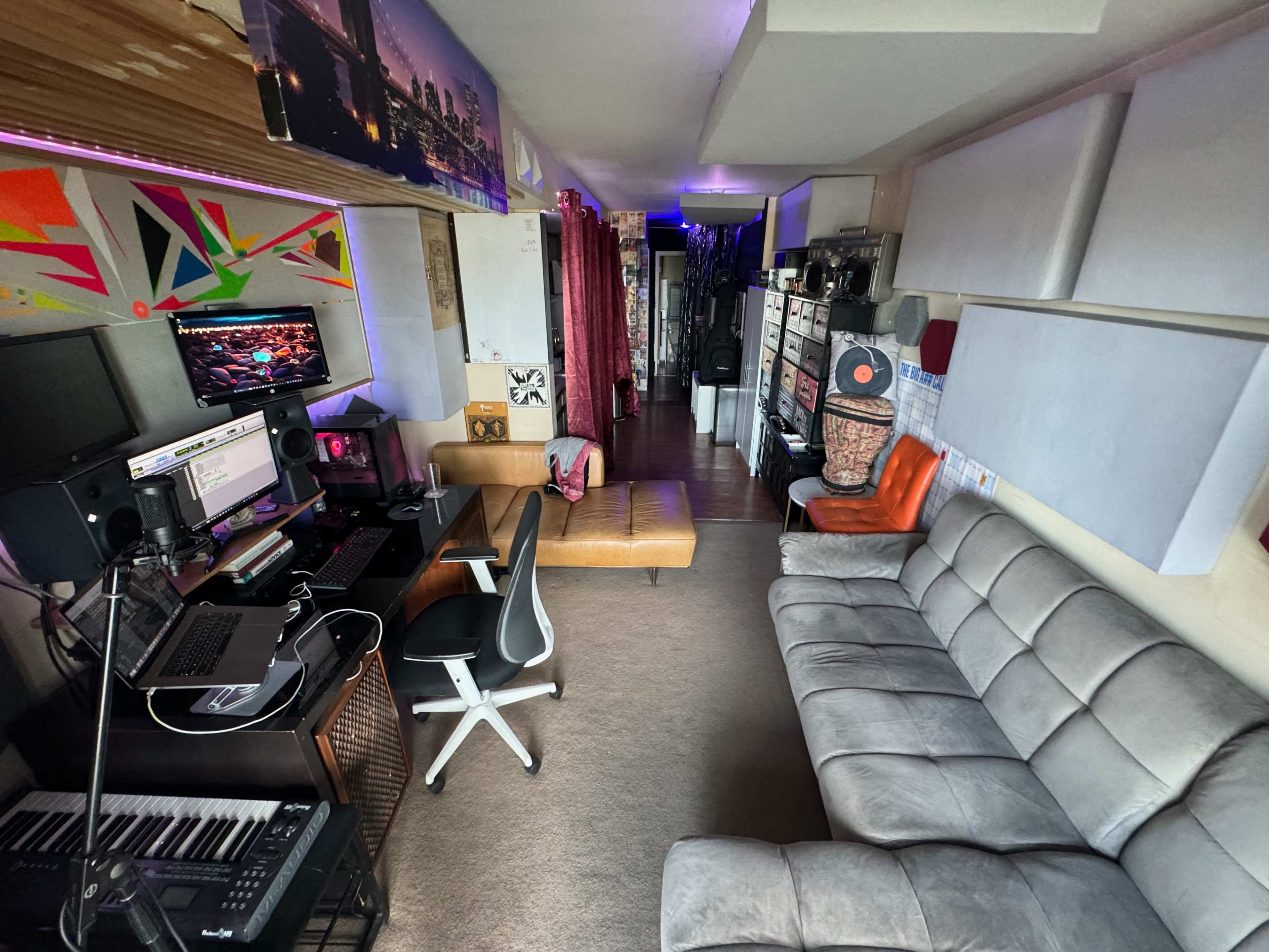 The image shows a narrow room featuring a workspace with a computer setup, a music keyboard, a gray sofa, an orange chair, and various decorative elements along the walls.