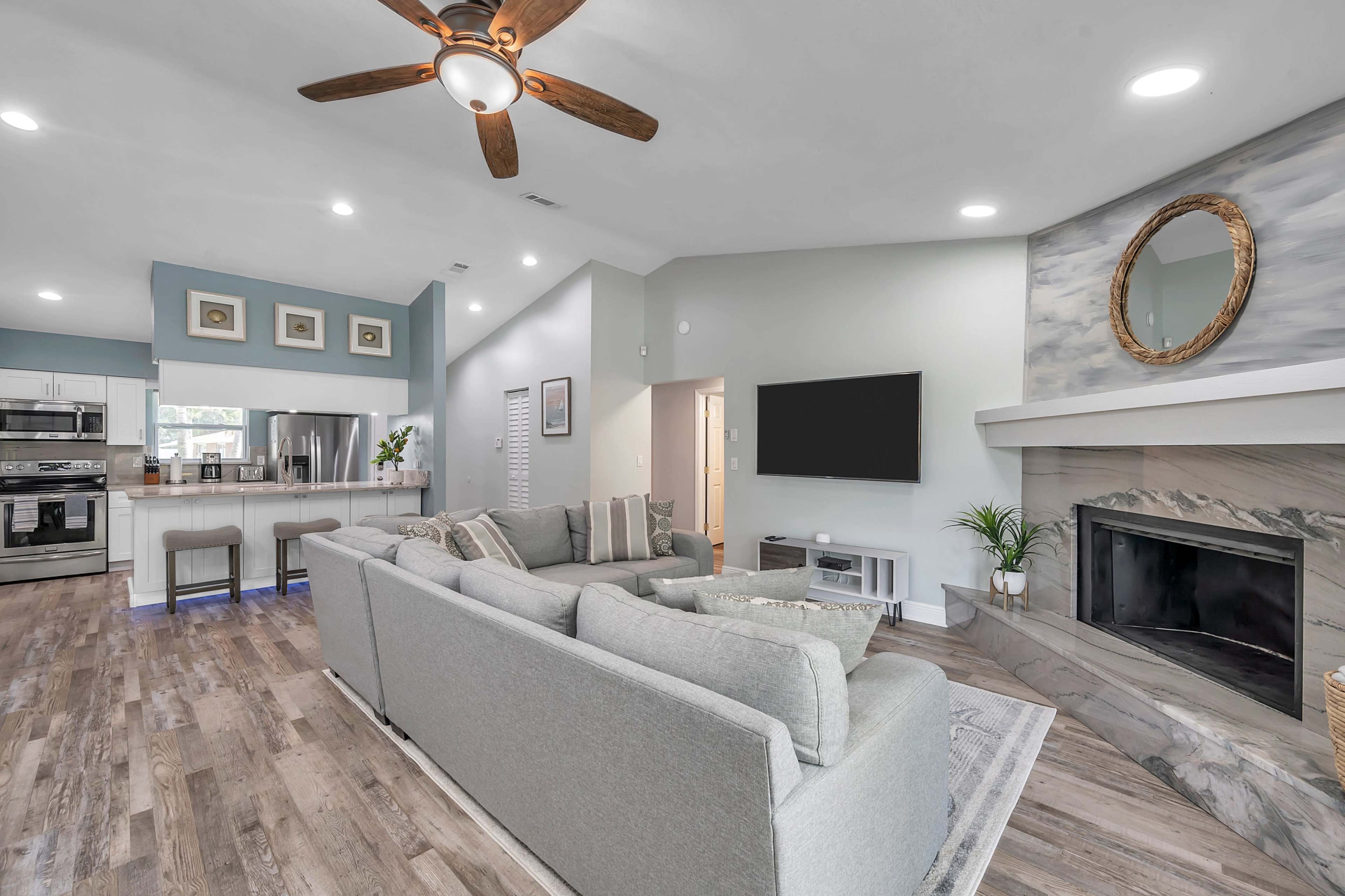 A modern living room features a comfortable light gray sectional sofa, a fireplace, and a wall-mounted television, with a ceiling fan and a well-lit open kitchen in the background.