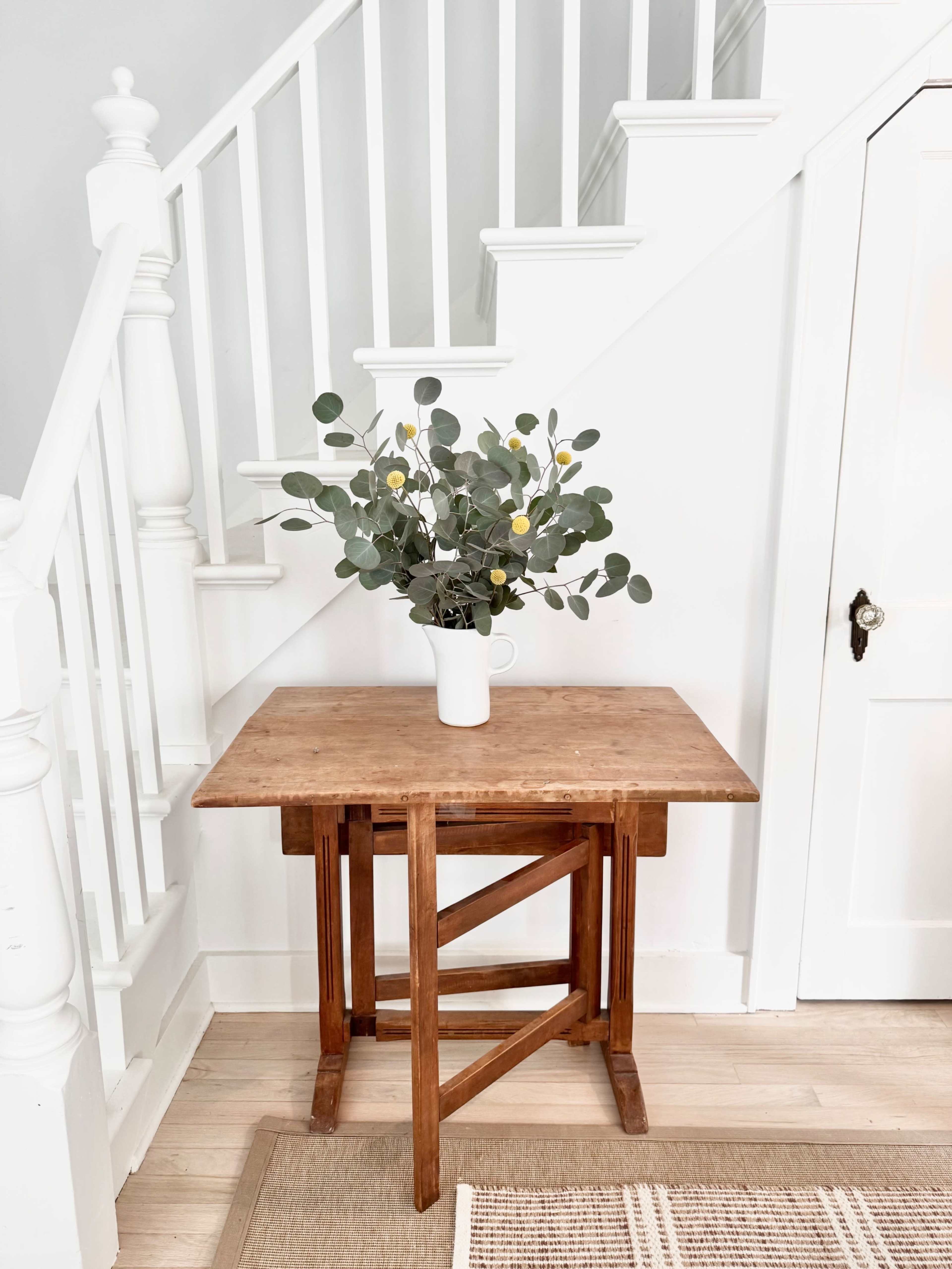 A wooden table with a vase of eucalyptus branches is positioned near a staircase in a bright entryway.