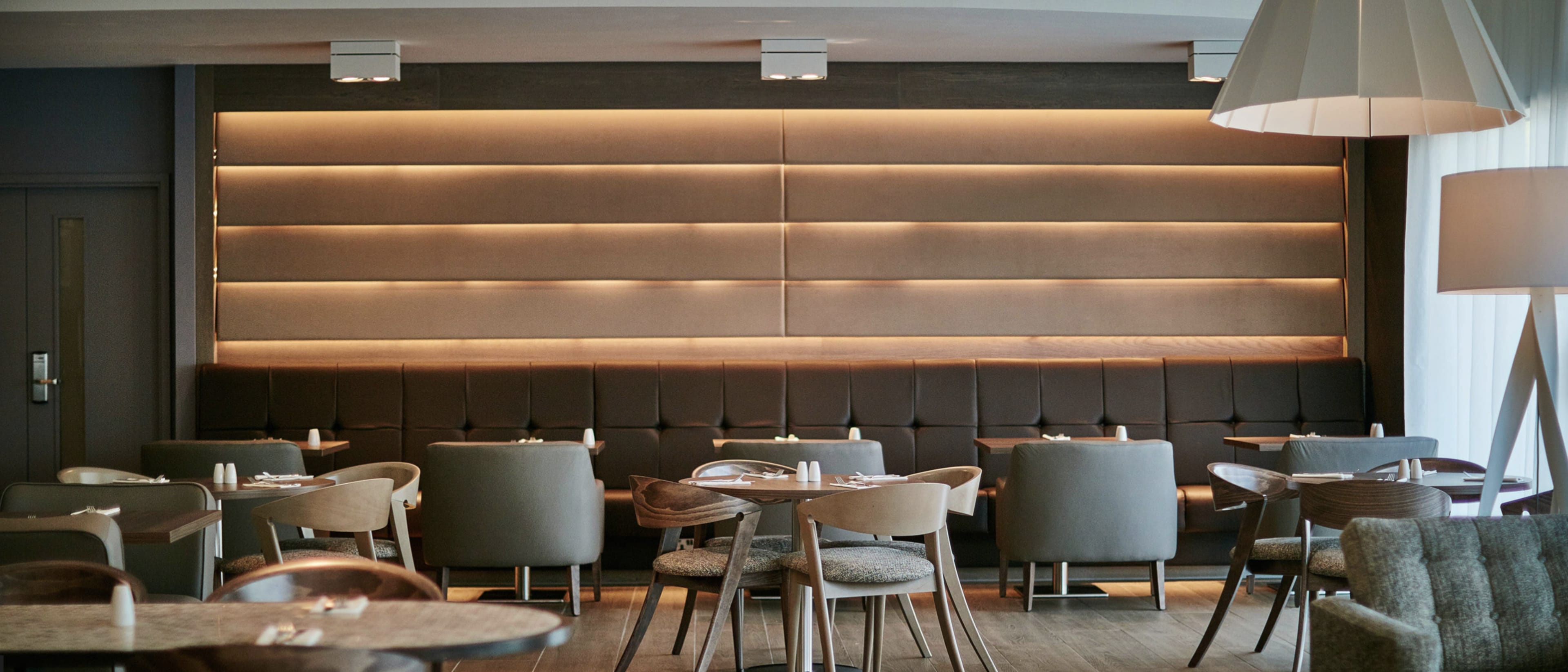 A modern restaurant interior features a series of wooden tables surrounded by various upholstered chairs, with a softly lit wall panel behind a sleek seating area.