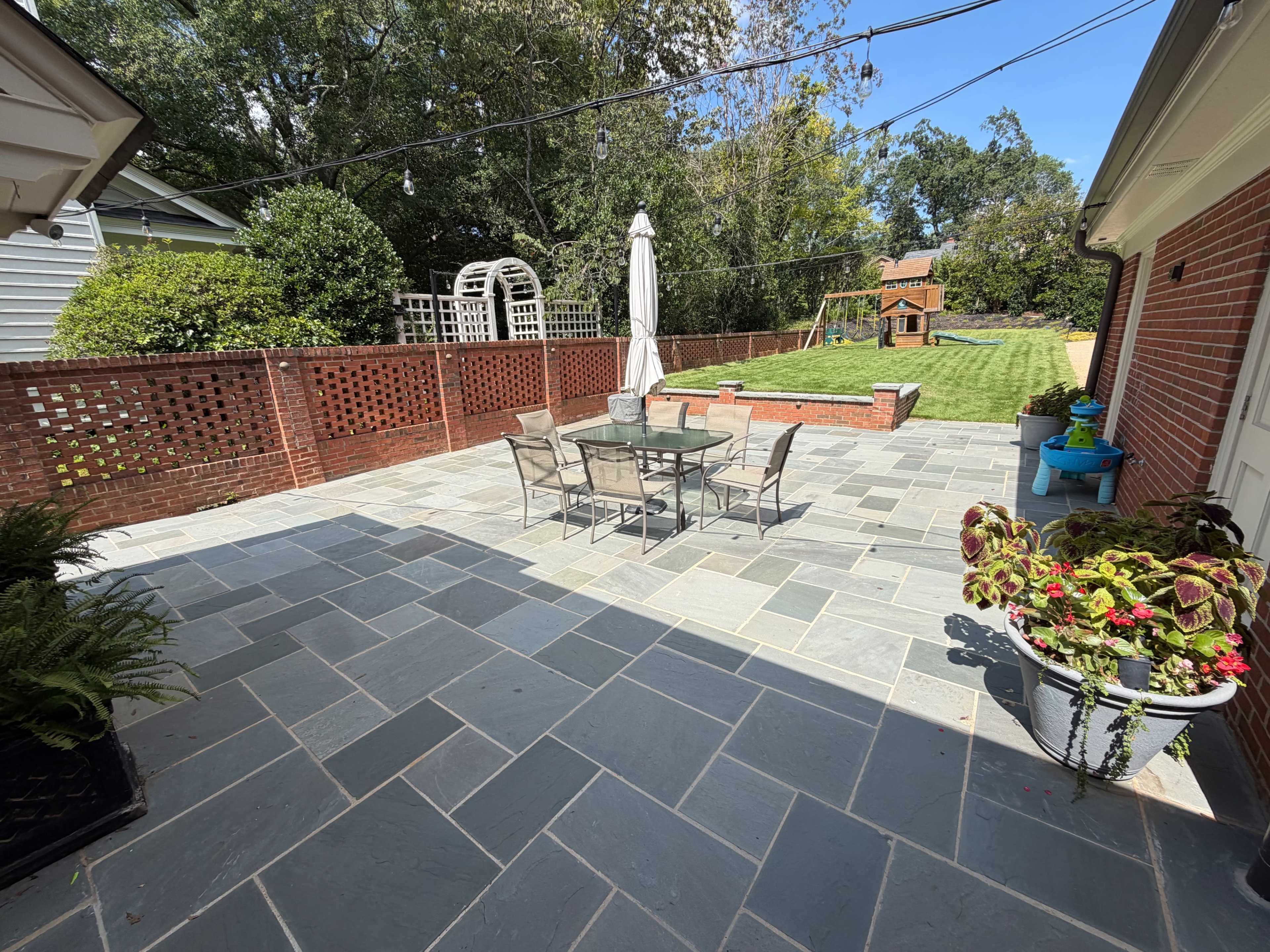 The image shows a patio area with a slate stone surface, a table and chairs, and a view of a well-maintained lawn and garden beyond a brick wall.