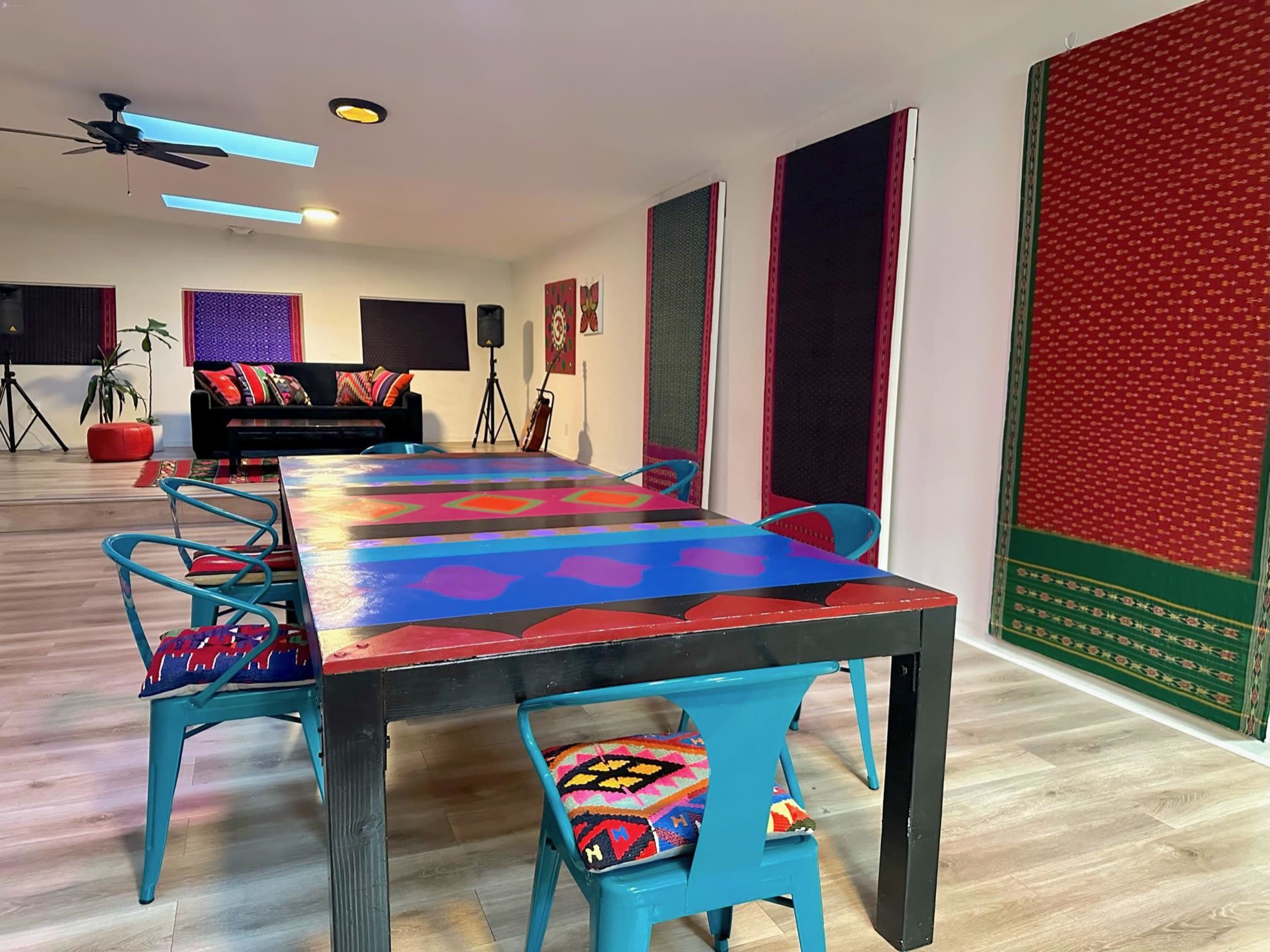 A spacious room featuring a large, colorful table surrounded by blue chairs, with decorated walls and a black sofa in the background.