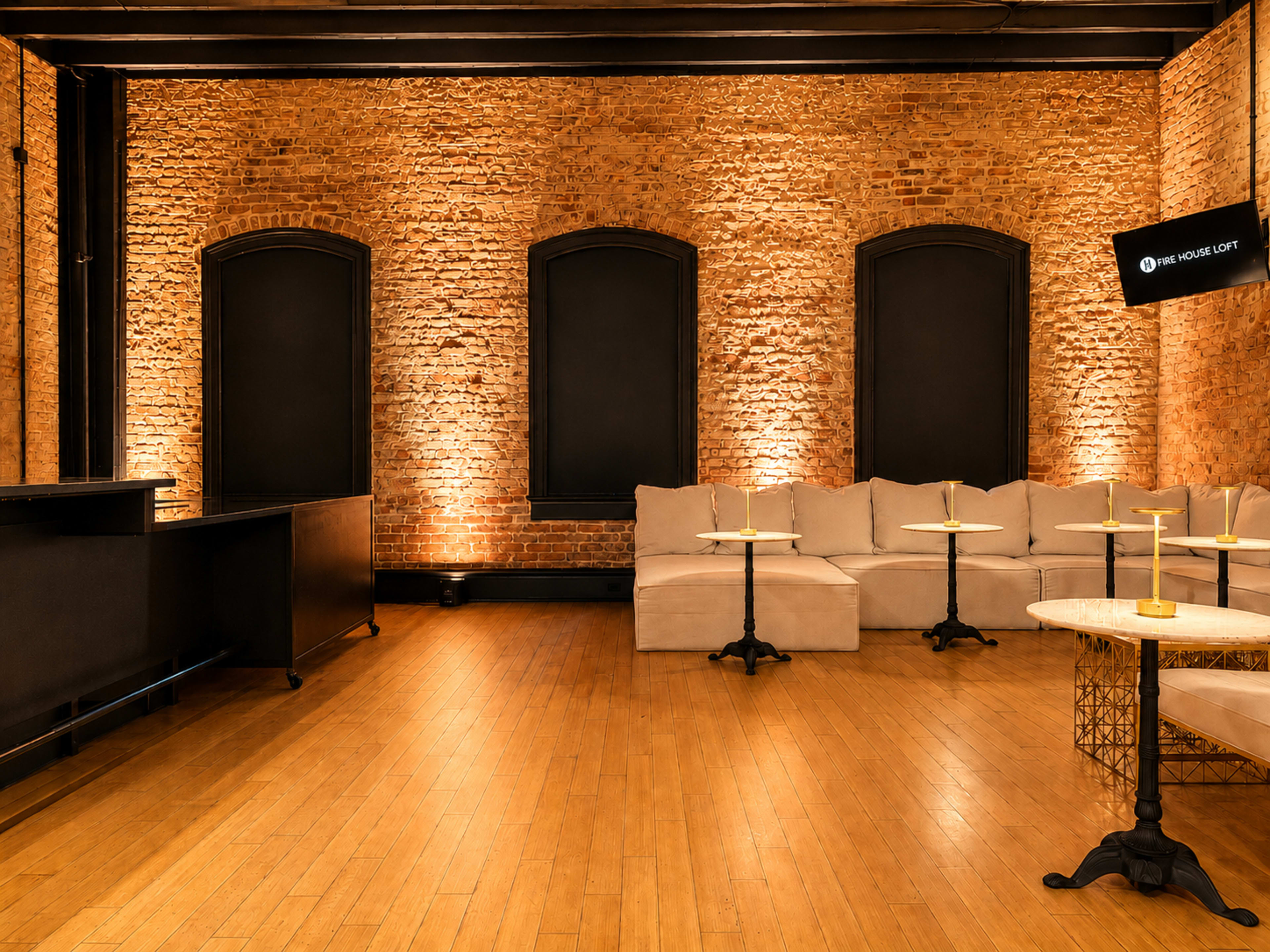 The image shows a spacious interior with exposed brick walls, two tall windows, and several light-colored couches arranged around small tables, illuminated by warm lighting.