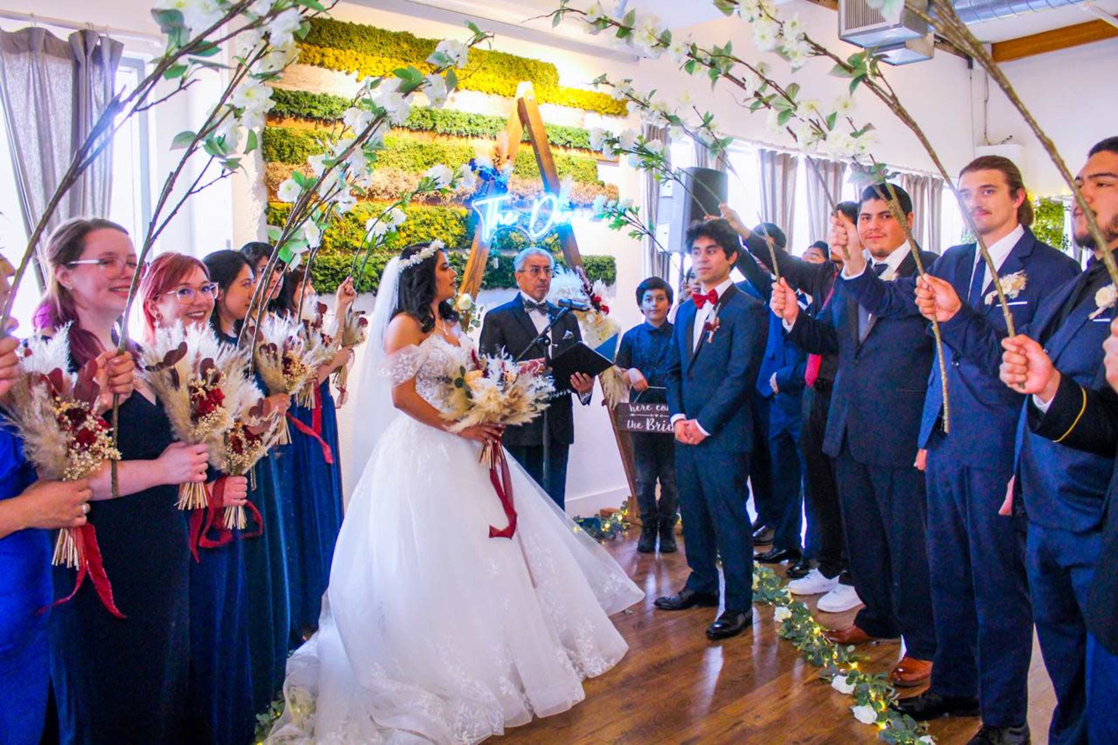 A bride and groom stand under an arch made of branches while surrounded by their wedding party in a decorated indoor setting.