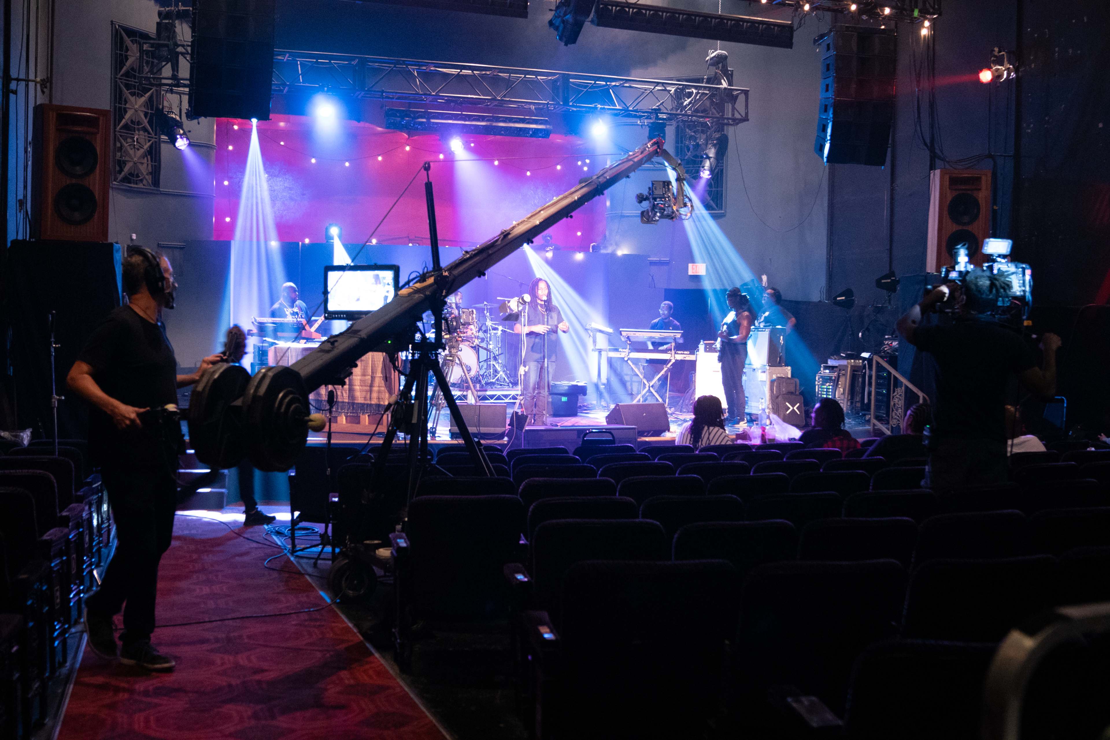 A live music performance is being filmed on stage, with crew members operating cameras and equipment amidst empty audience seats.