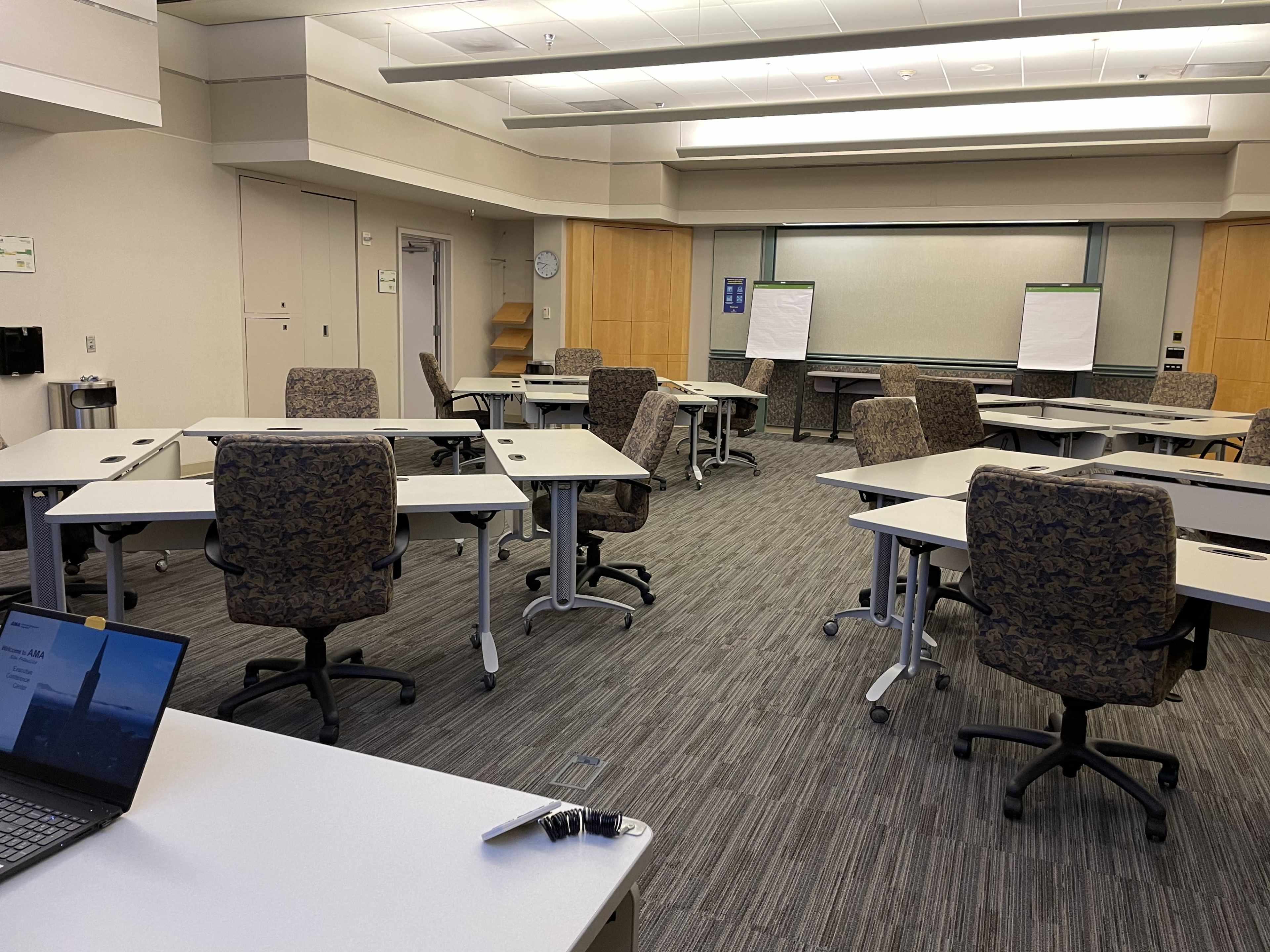A conference room with several tables and chairs arranged for a meeting, featuring a laptop on a table in the foreground.