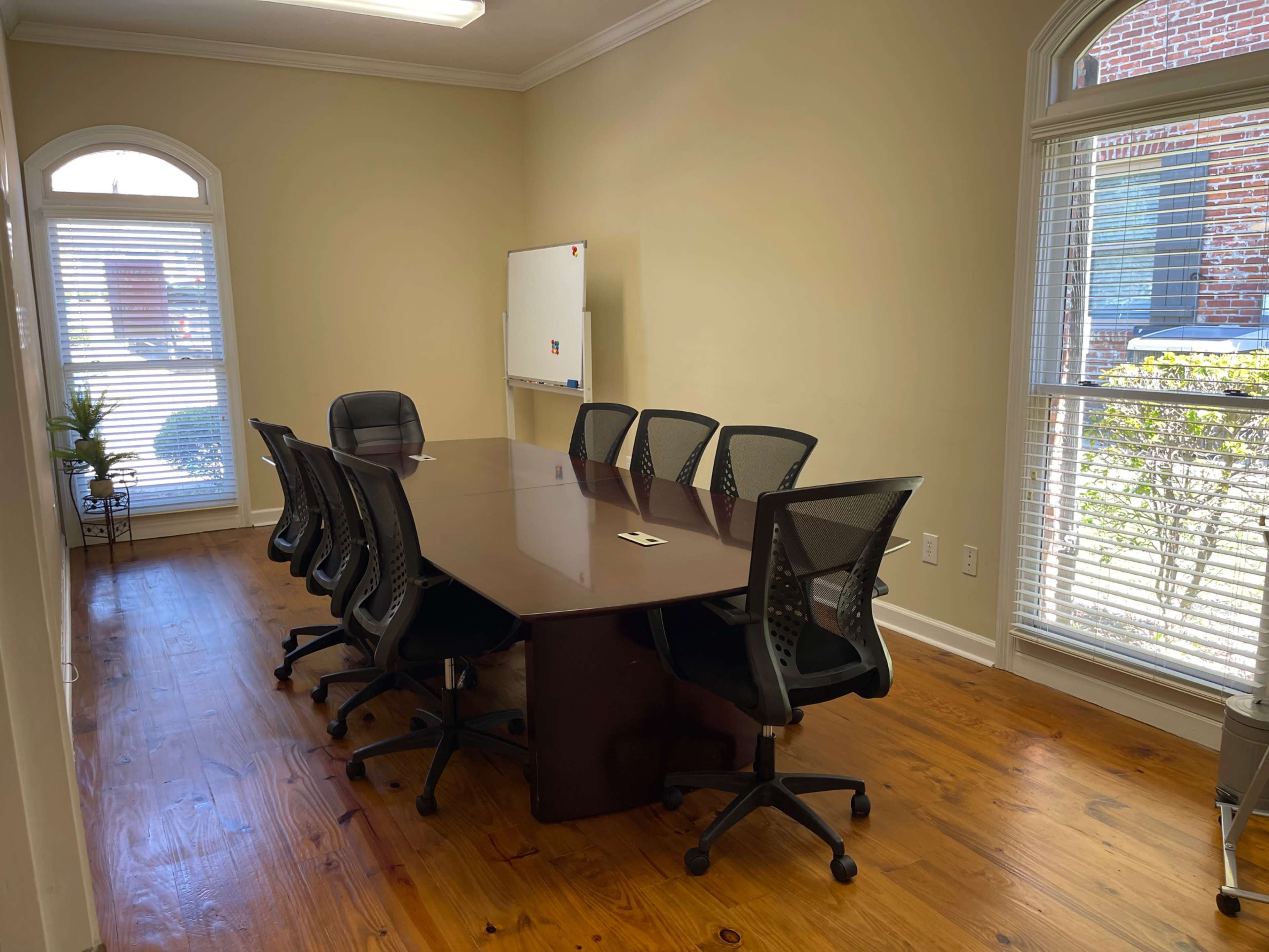 A conference room features a large wooden table surrounded by eight ergonomic chairs, with a whiteboard on the wall and windows letting in natural light.