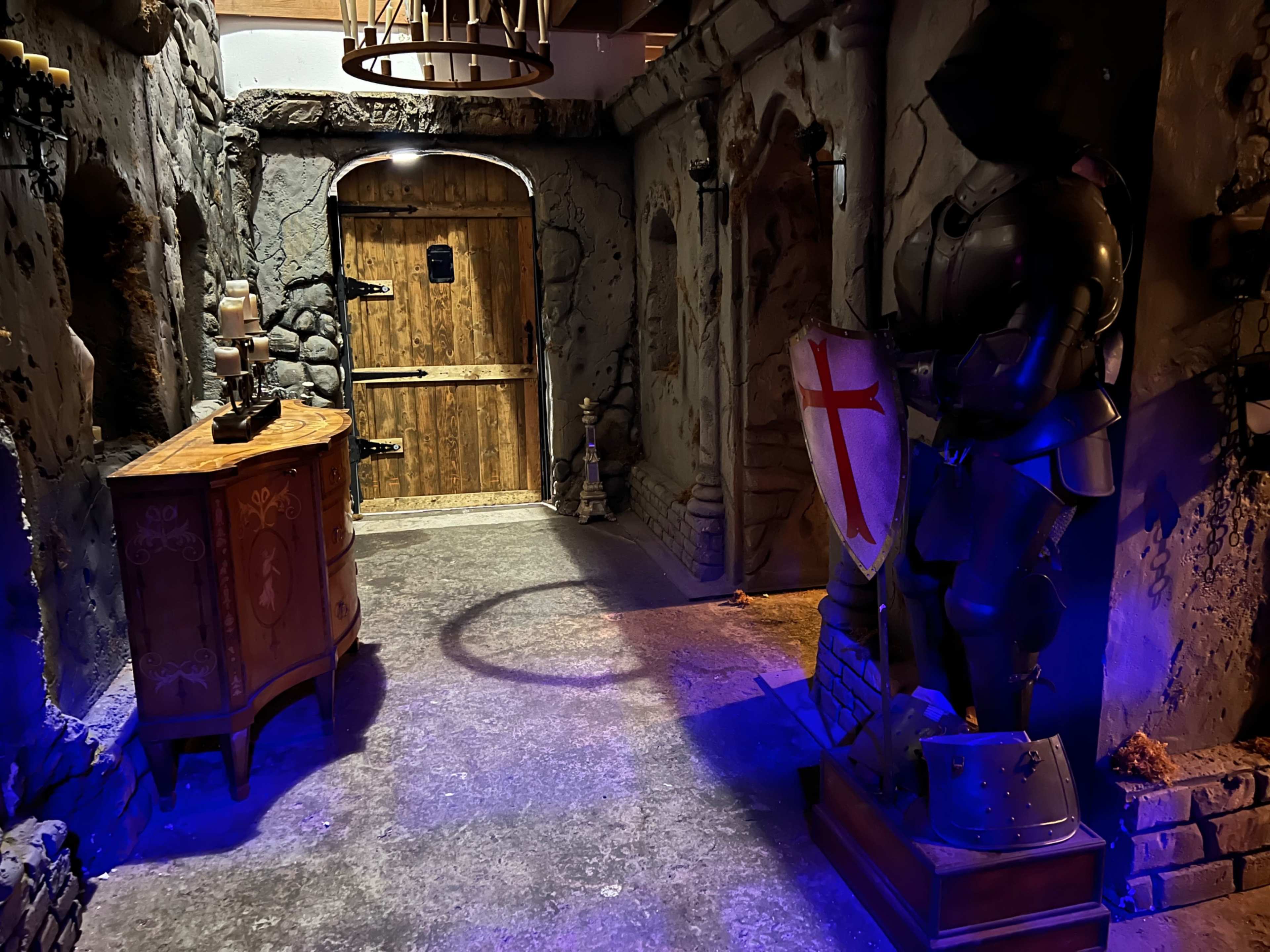 The image depicts a stone-walled corridor with a wooden door at the end, flanked by a suit of armor and a wooden cabinet.
