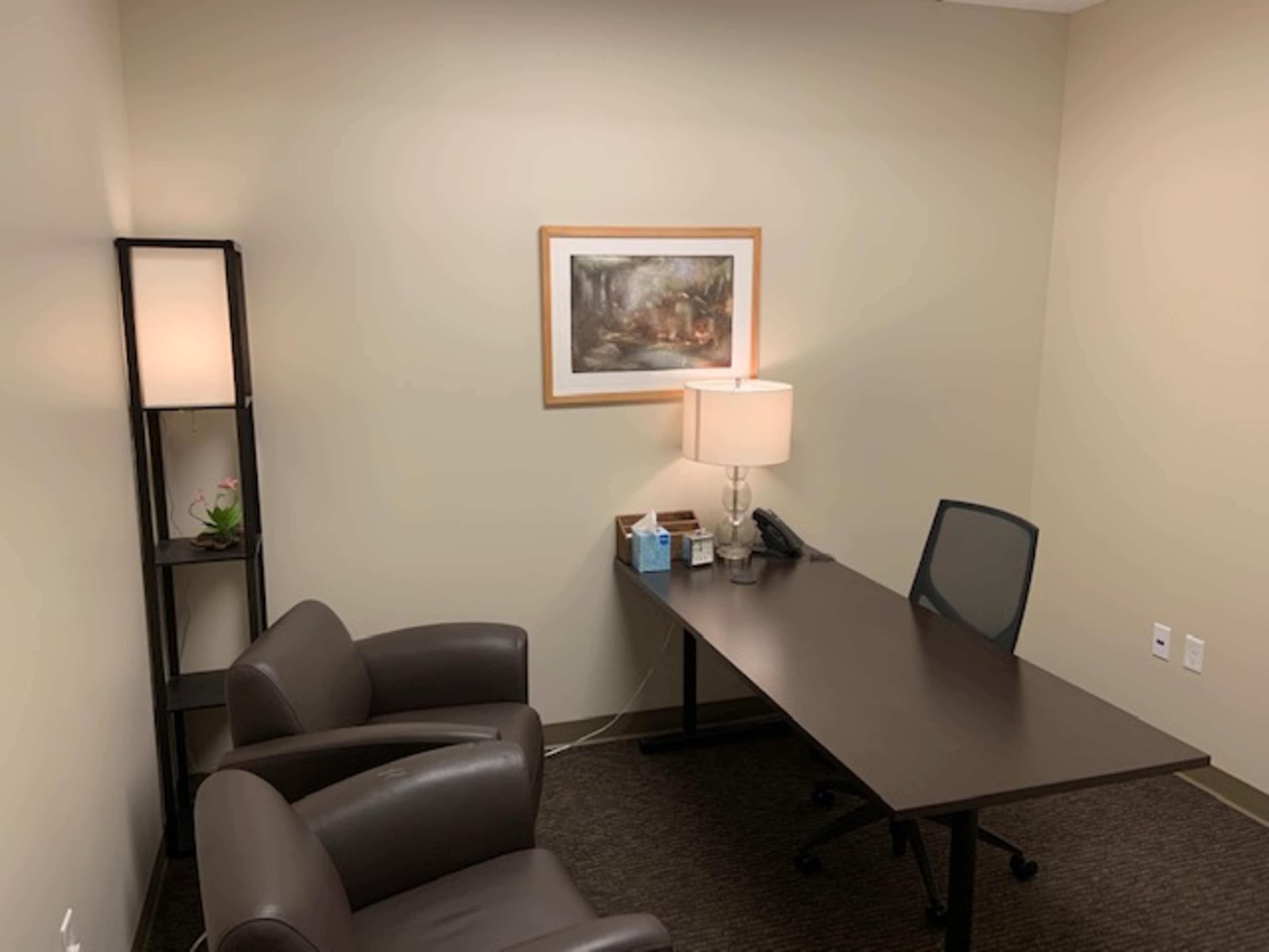 A small office with a desk, a chair, a lamp, and a framed artwork on the wall.