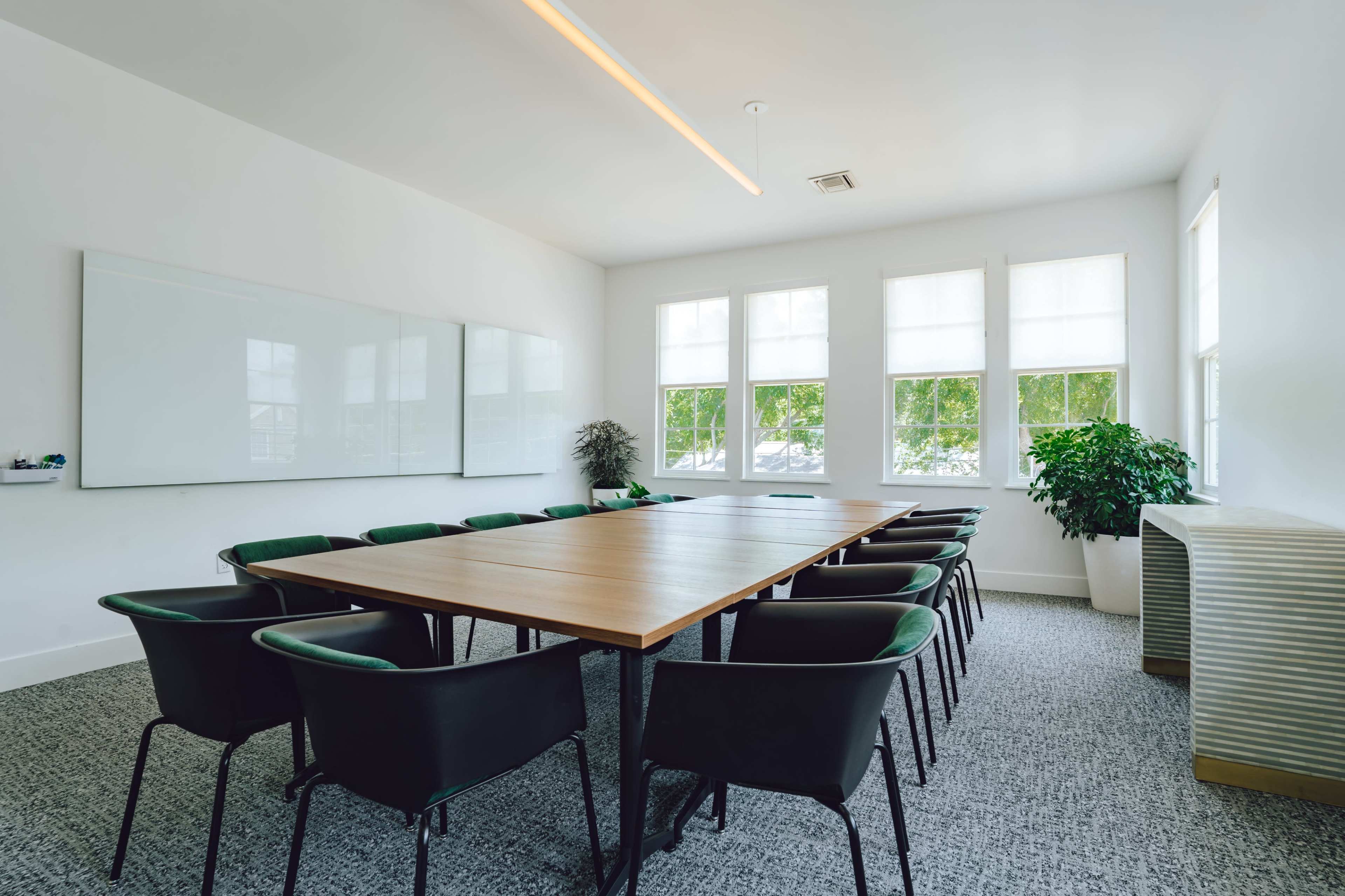 The image shows a modern conference room with a large wooden table surrounded by black chairs, large windows letting in natural light, and whiteboards on the wall.
