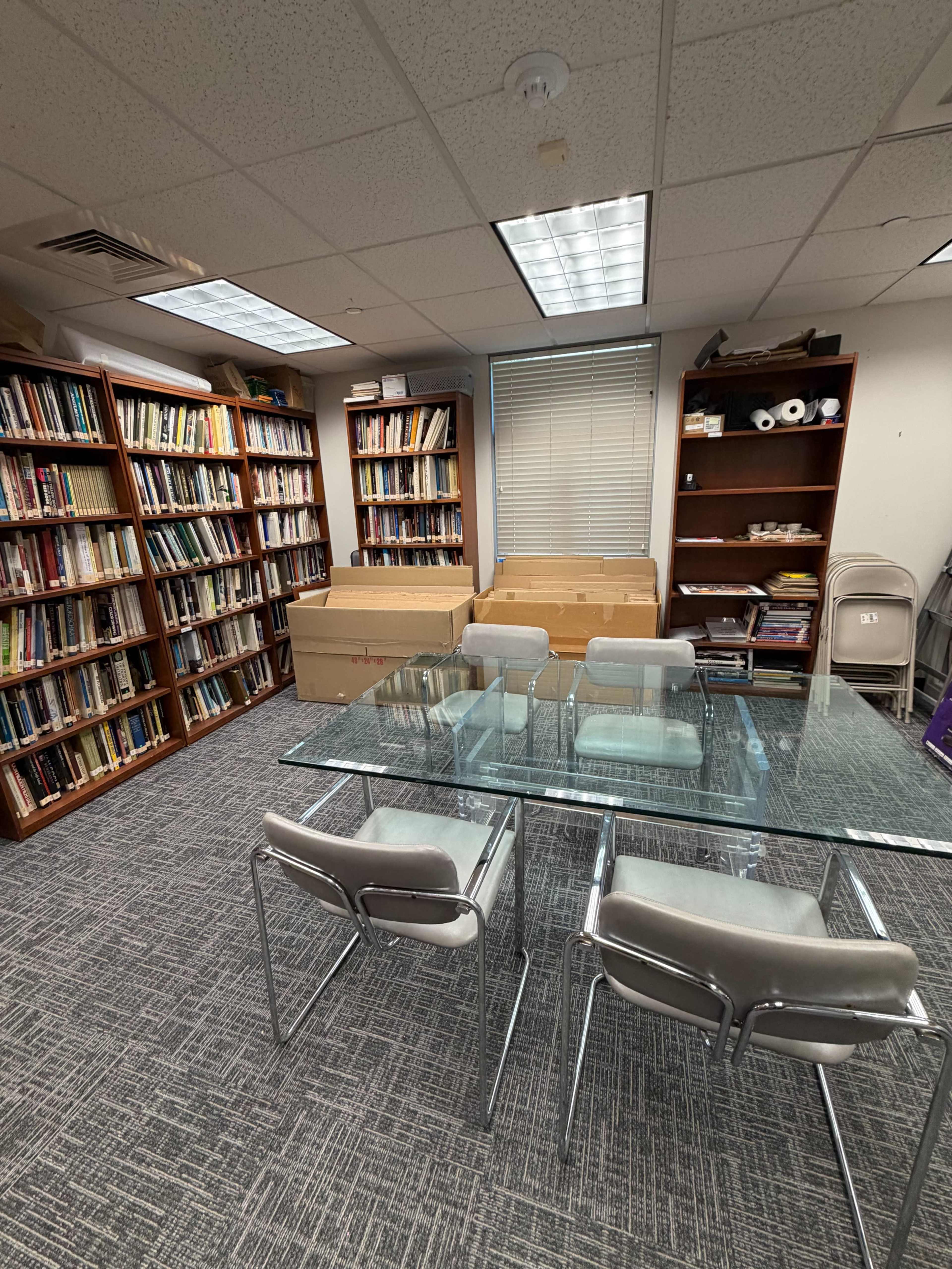 The image shows a small office with bookshelves filled with books, a glass table surrounded by chairs, and several cardboard boxes stacked in one corner.