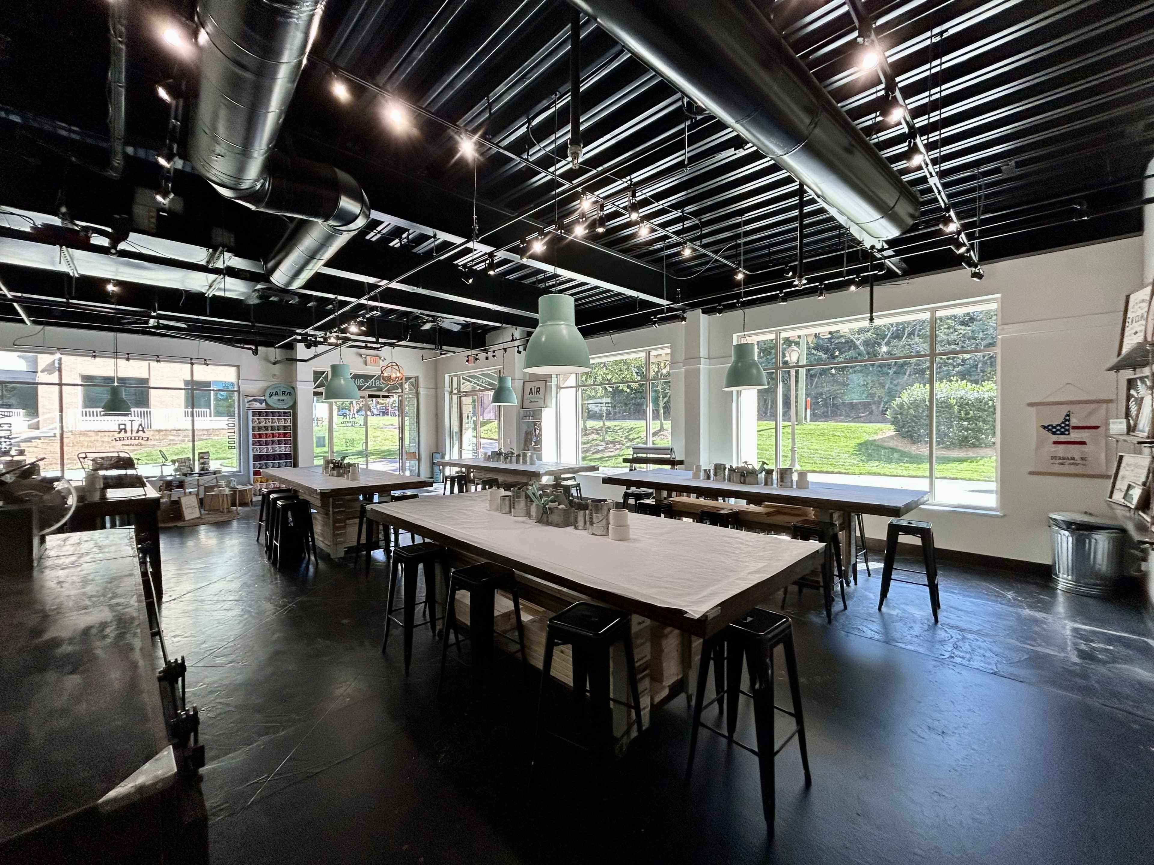 A spacious room with long tables and industrial-style lighting, featuring large windows that overlook a green outdoor area.