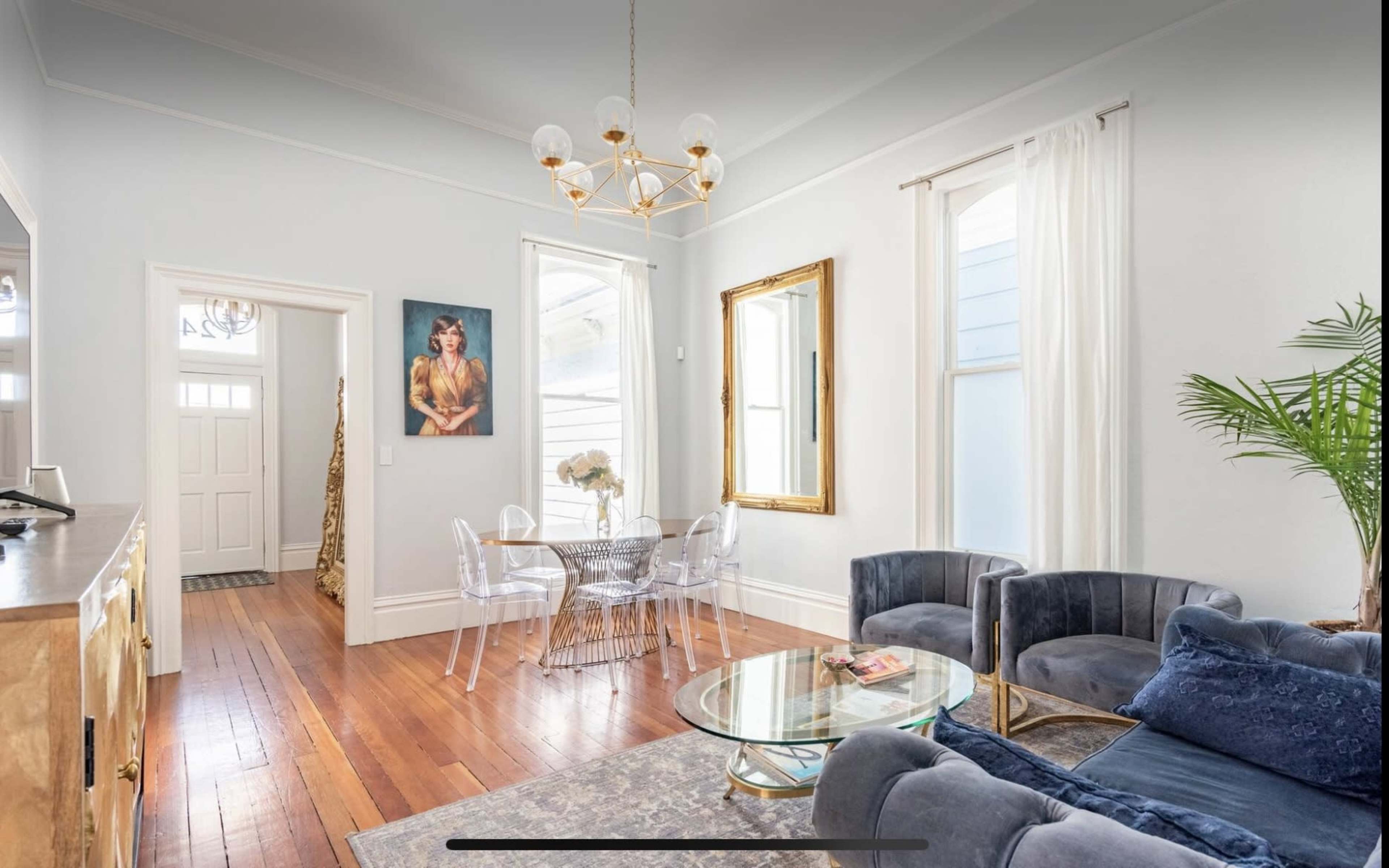 The room features a modern living area with a circular glass table surrounded by transparent chairs, a plush blue sofa, and a large mirror, complemented by natural light coming through two windows.