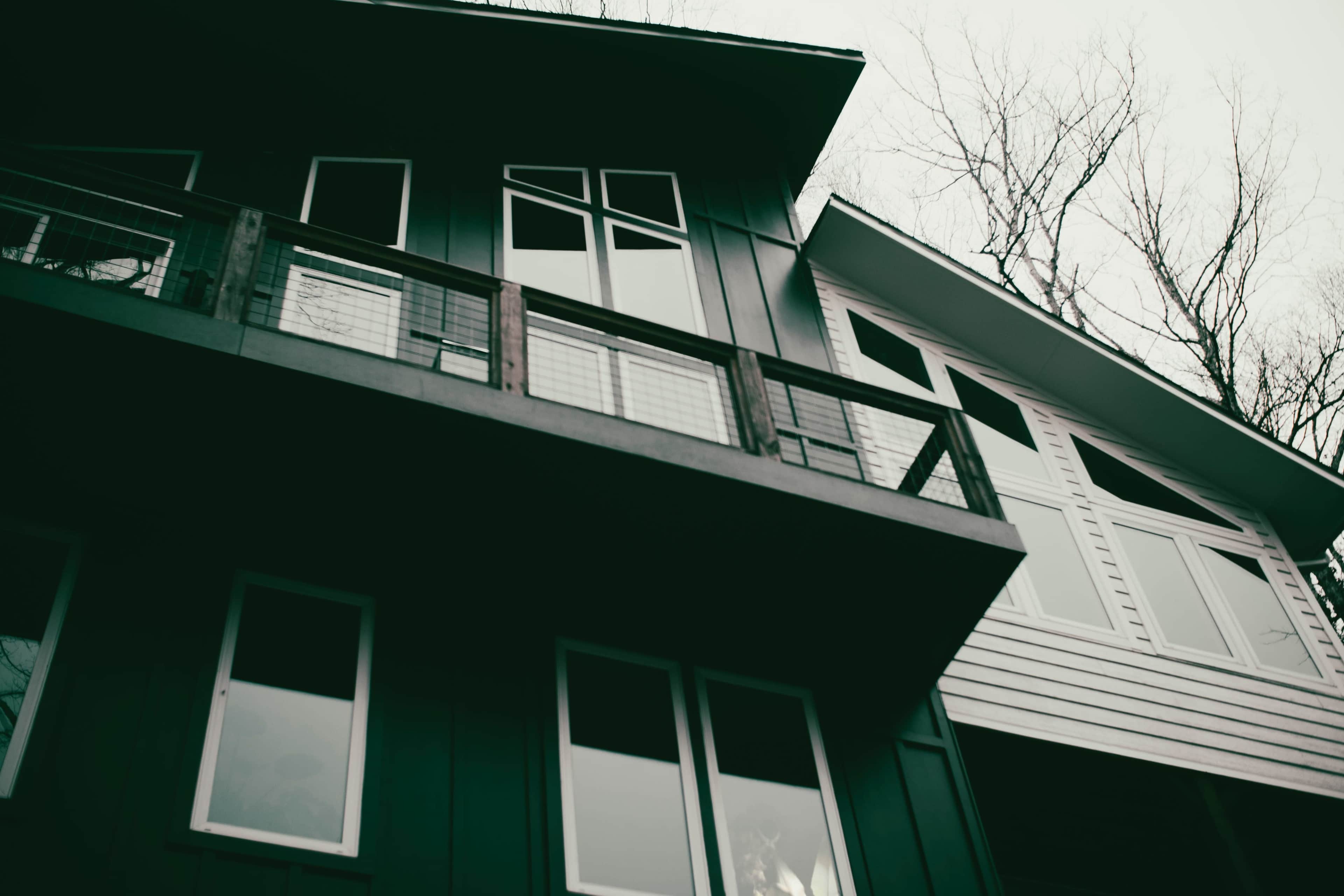 The image shows a modern house with multiple large windows and a balcony, set against a backdrop of bare trees.
