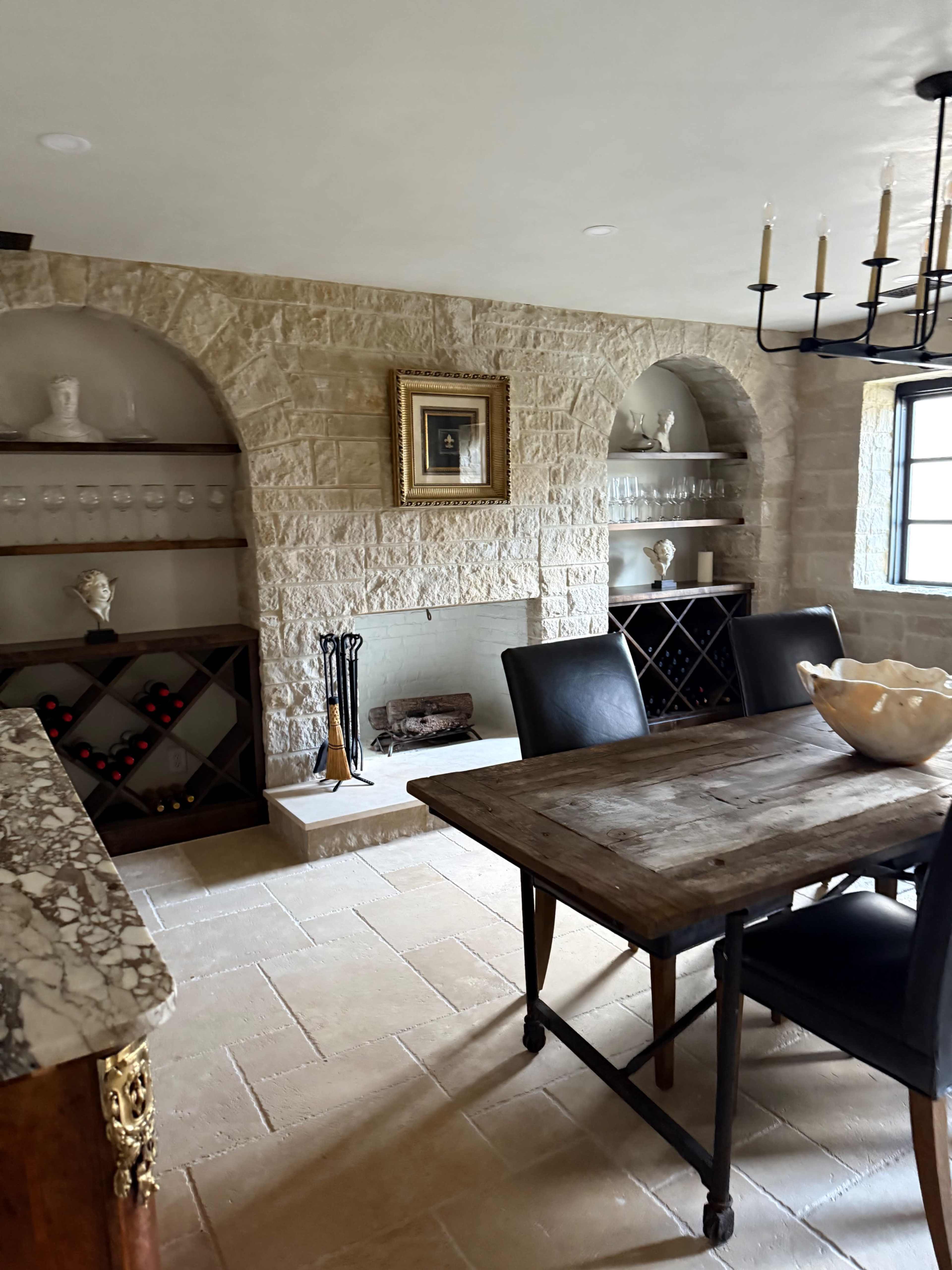 The image shows a rustic dining area featuring a wooden table, black chairs, a stone fireplace with decorative shelves, and a wine rack.