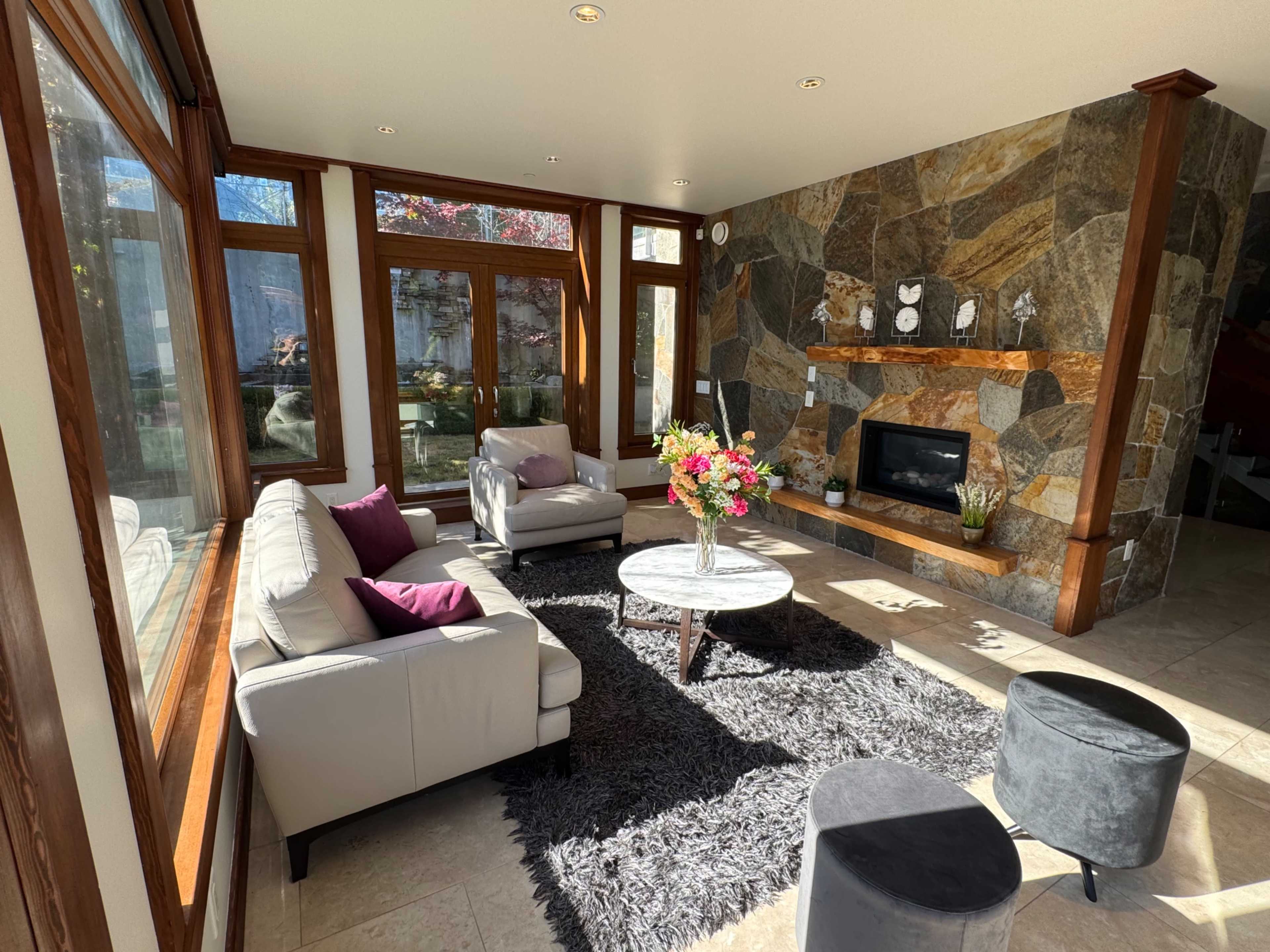 The image shows a modern living room with two sofas, a coffee table, a stone fireplace, and natural light coming through large windows.