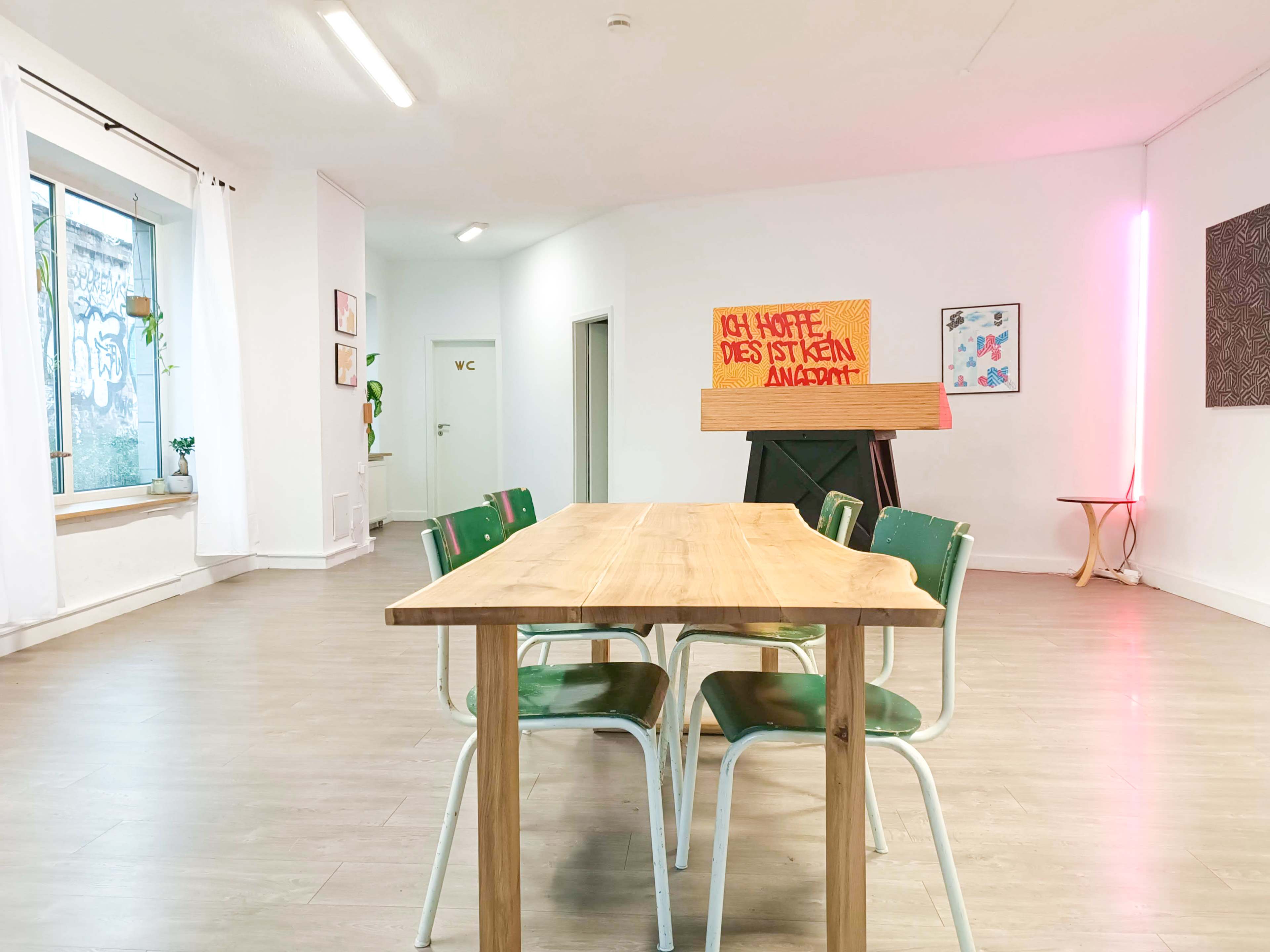 A wooden table with green chairs is positioned in a bright, minimalist room featuring white walls and various art pieces.