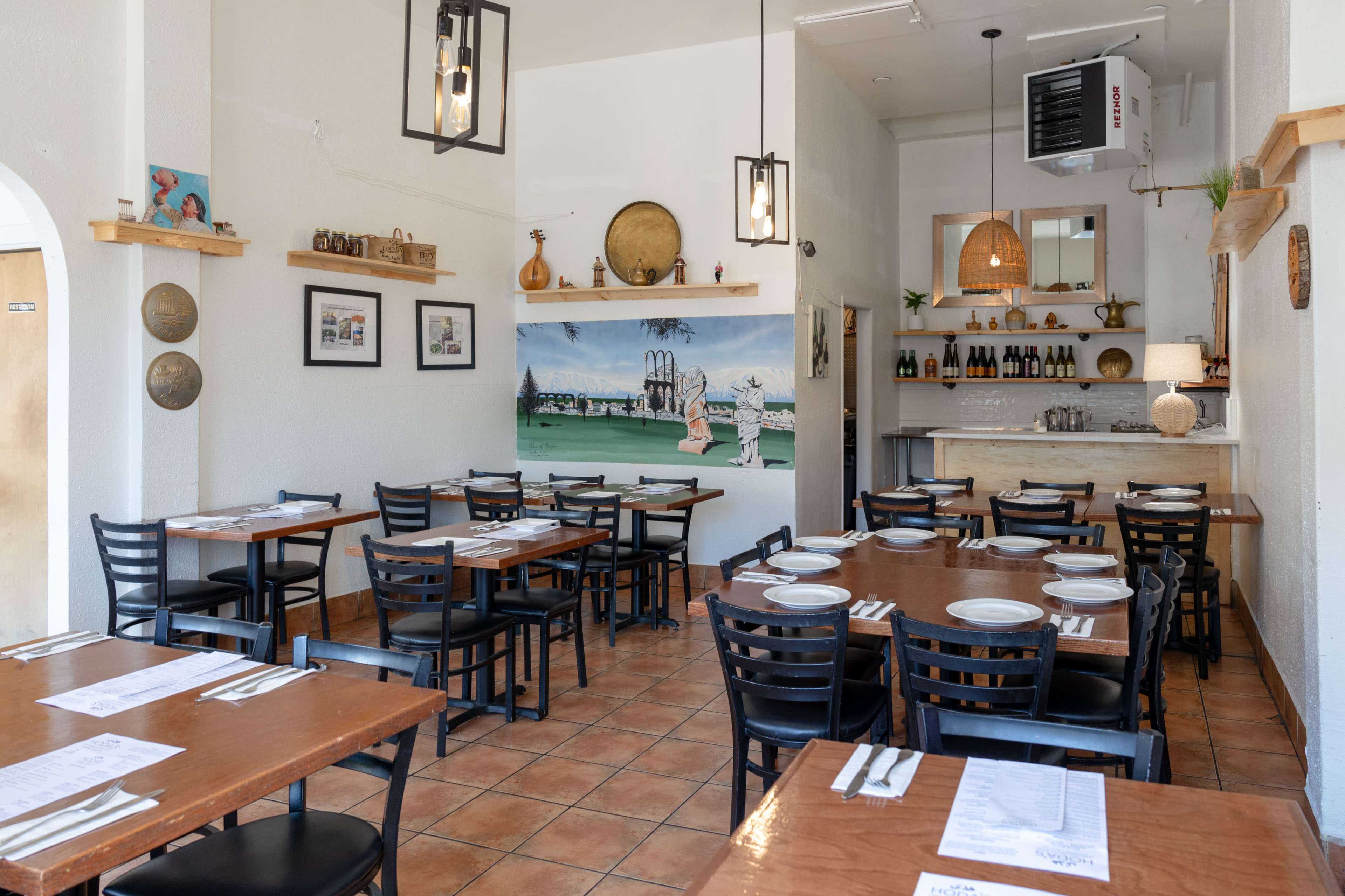 The interior of a restaurant features wooden tables and chairs, art on the walls, and a serving area with a countertop and shelves.